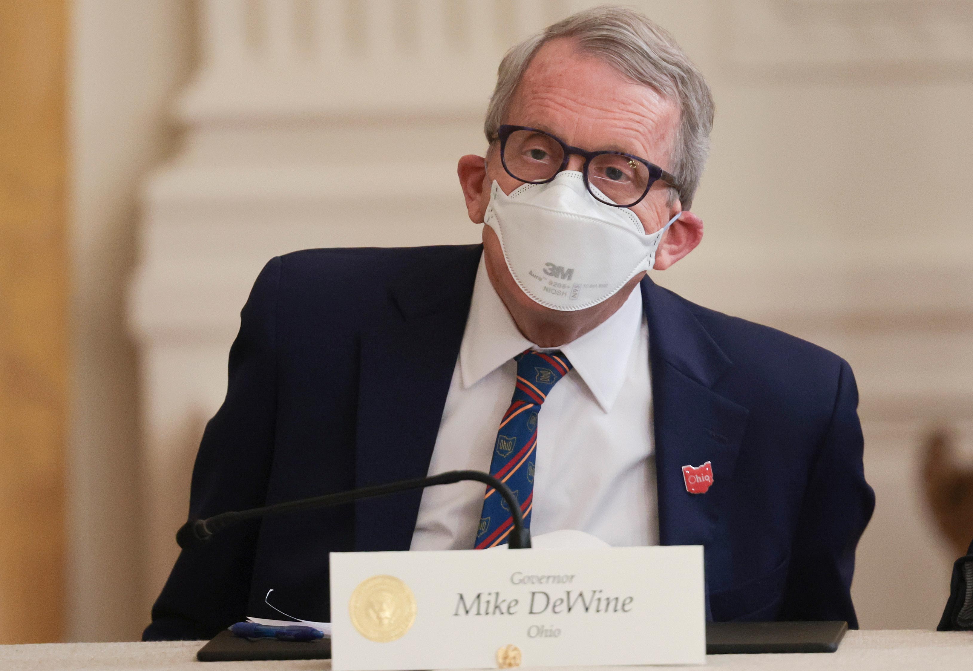 A photo shows Gov. Mike DeWine sitting at a table with a name card in front of him.