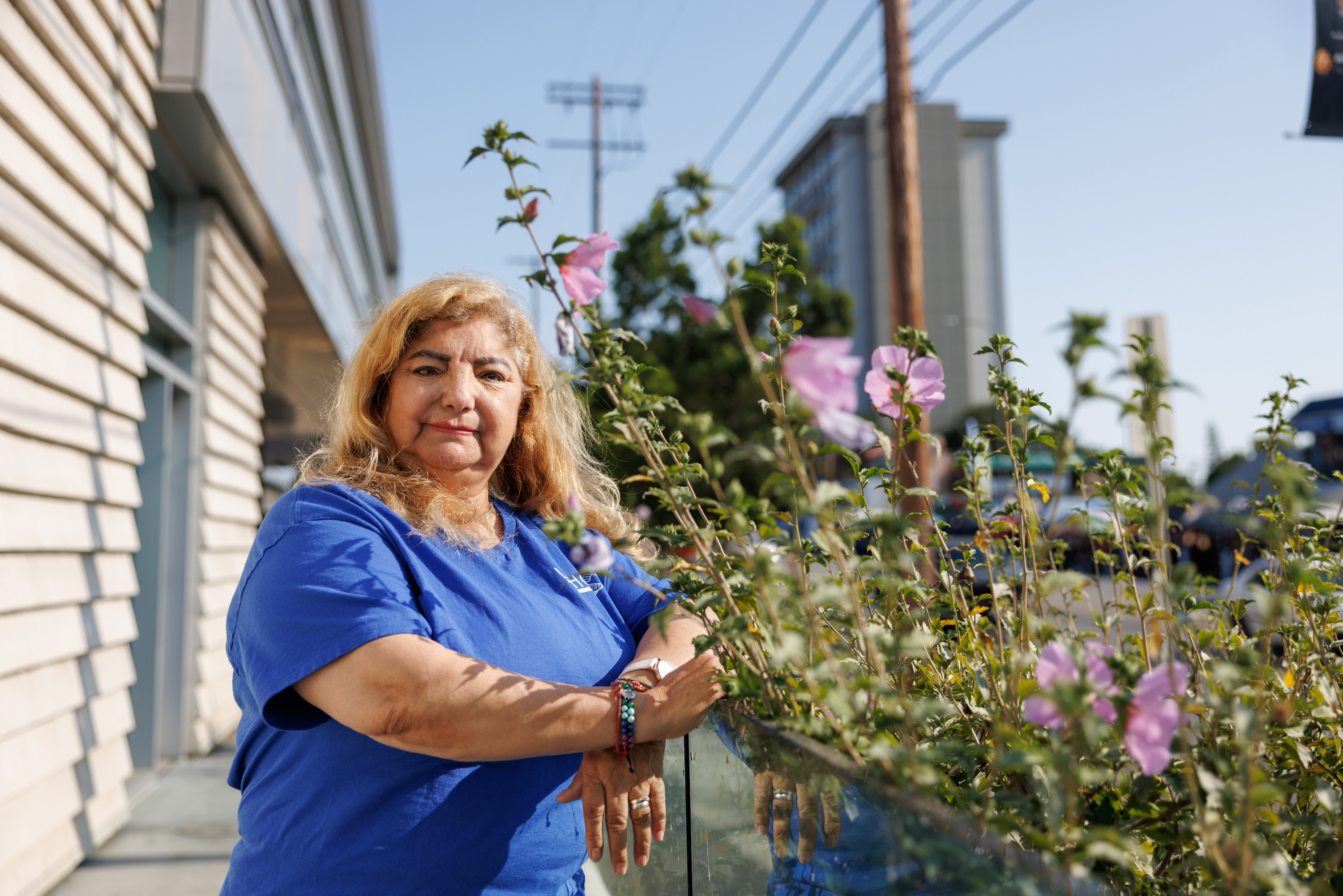 A woman wearing a blue t-shirt stands next to a pink flowering bush as she looks at the camera