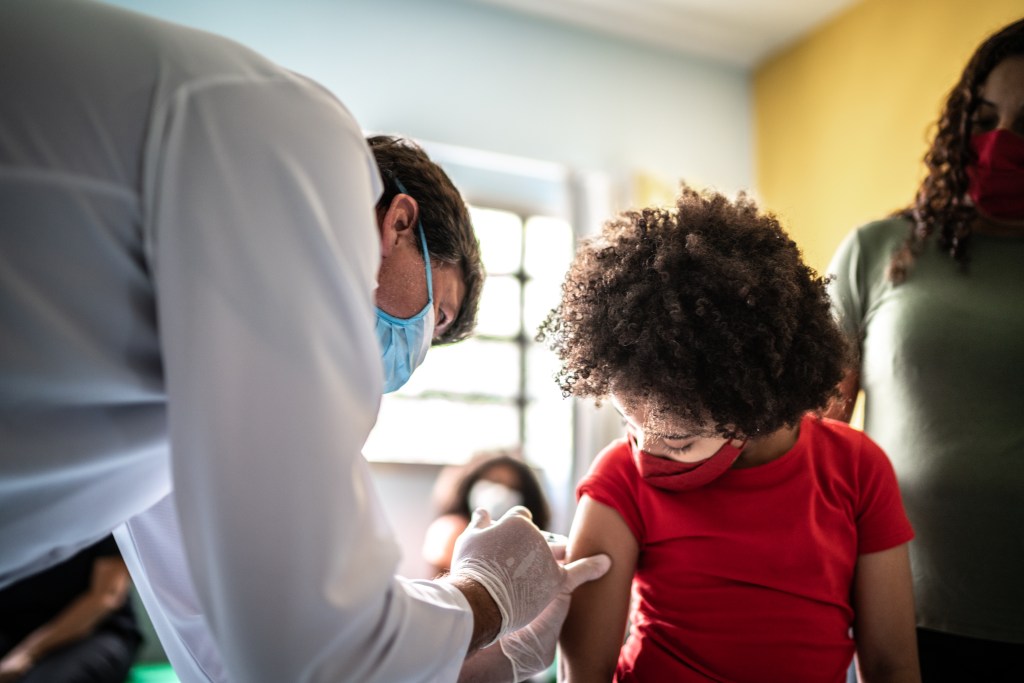 A child receives a medical shot in their arm from a doctor while their parent watches. All figures are masked.