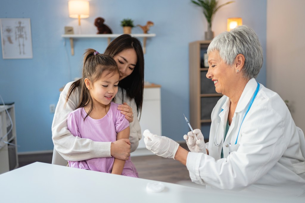 A Japanese child sits in her mother's lap while receiving a vaccine from a senior female Caucasian pediatrician.