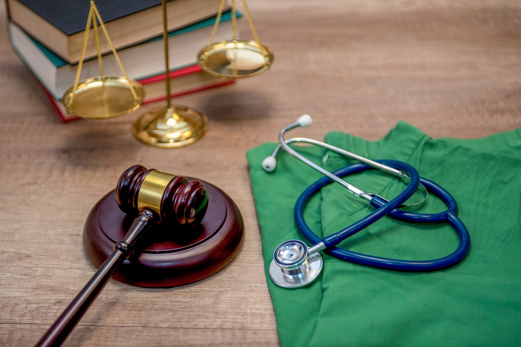 A gavel lays on a table next to a stethoscope on scrubs with a scale and a book stack in the background.