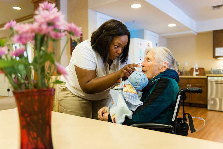 Resident Marilou Roos holds a baby doll while Jessica Butler, a life enrichment manager at Sunrise Senior Living in Beverly Hills, California, helps her take a drink of water on August 2, 2016. (Heidi de Marco/KHN)