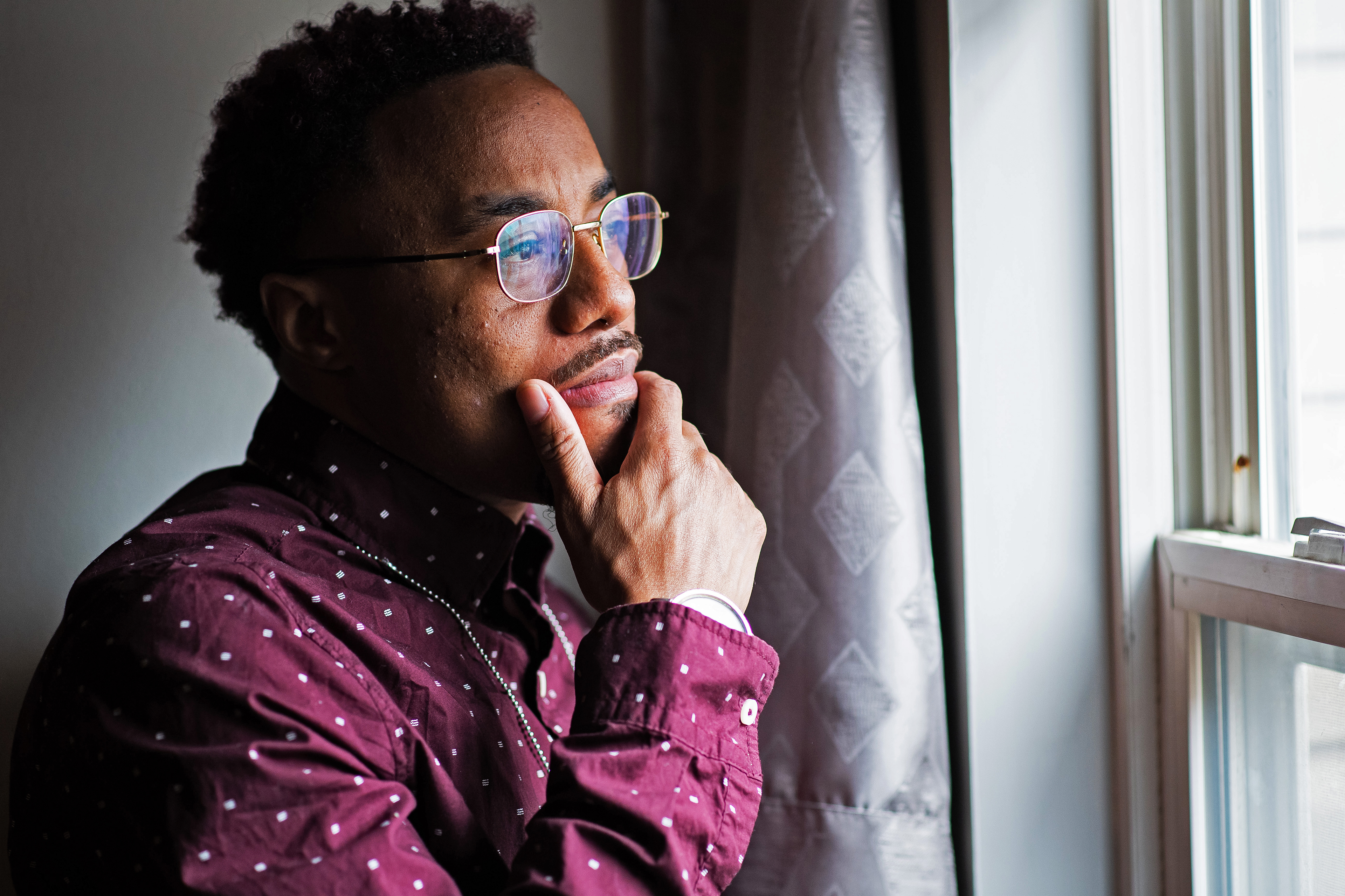 Nicholas Kelly is seen on the left looking out a window, holding his hand to his chin. Bright daylight shines in from the window, casting dramatic shadows on the left side of his body.