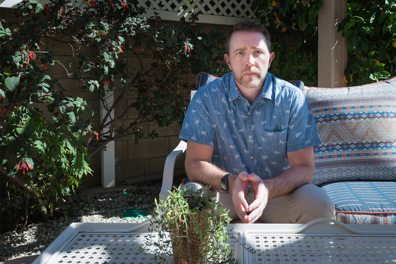 Lee Stonum sits on a deck chair outside of his home. The right side of his face and parts of a fence behind him are hit with dappled sunlight. He holds his hands together in his lap, looking forward at the camera.