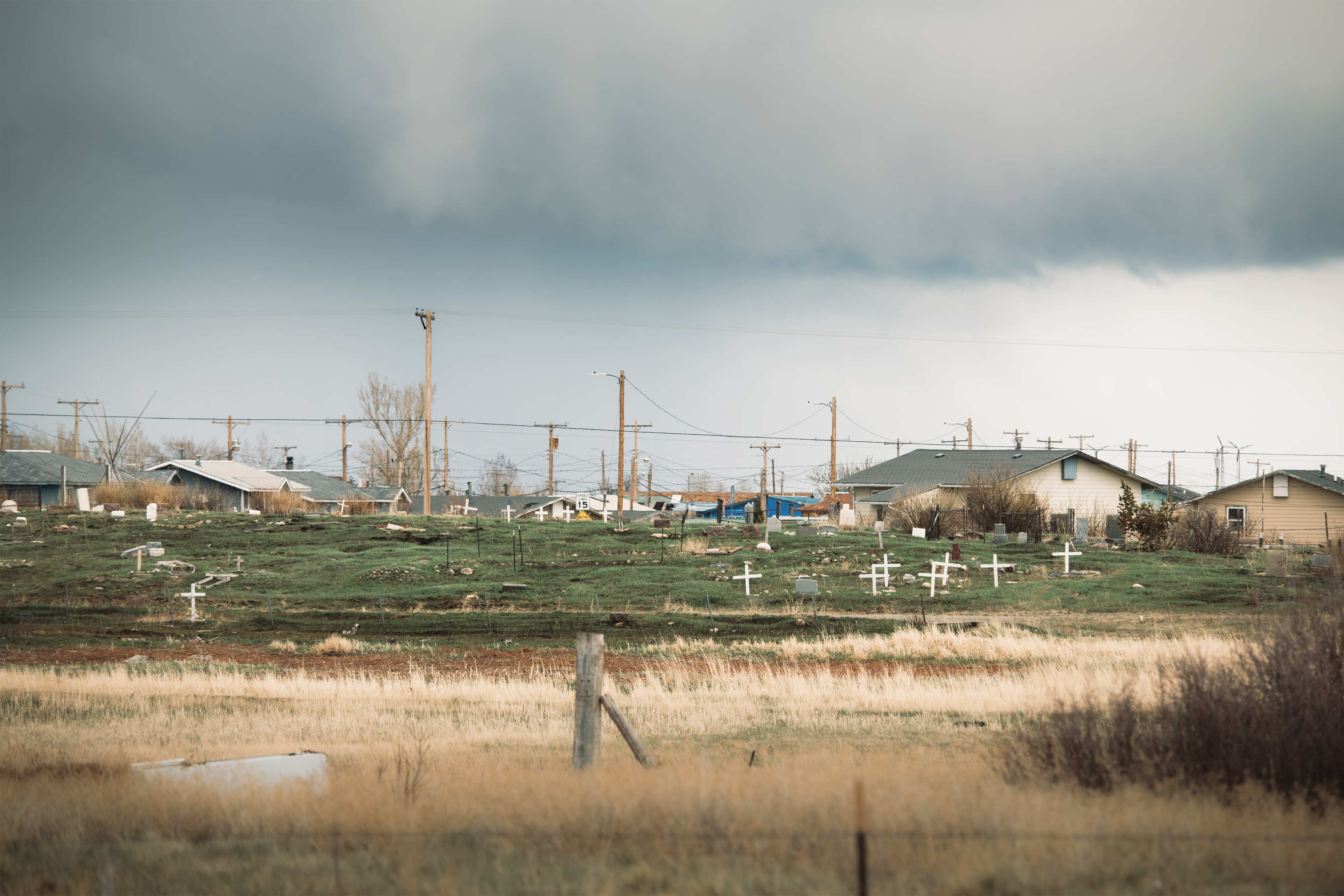 A photo shows a grassy cemetery from afar. Rows of houses are seen behind it.
