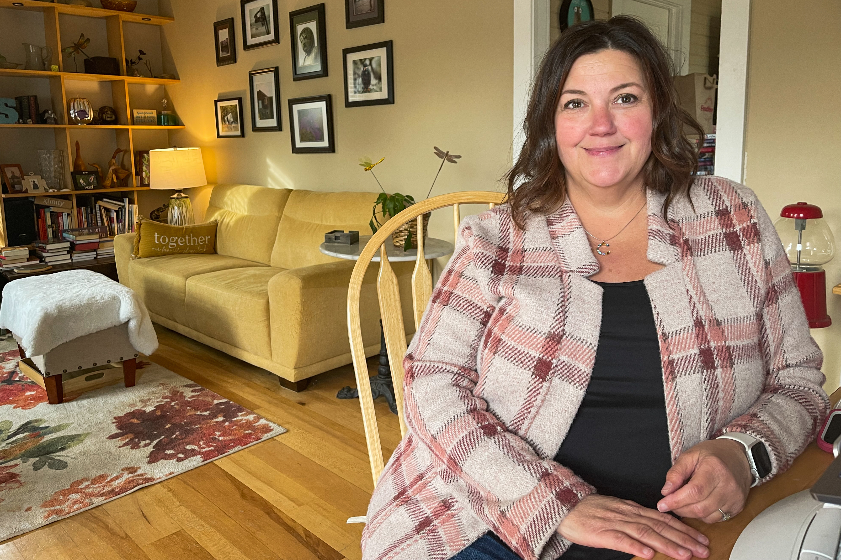A photo shows Cindy Stockton sitting in a chair inside her home. A sofa and a wall filled with photos is seen behind her.