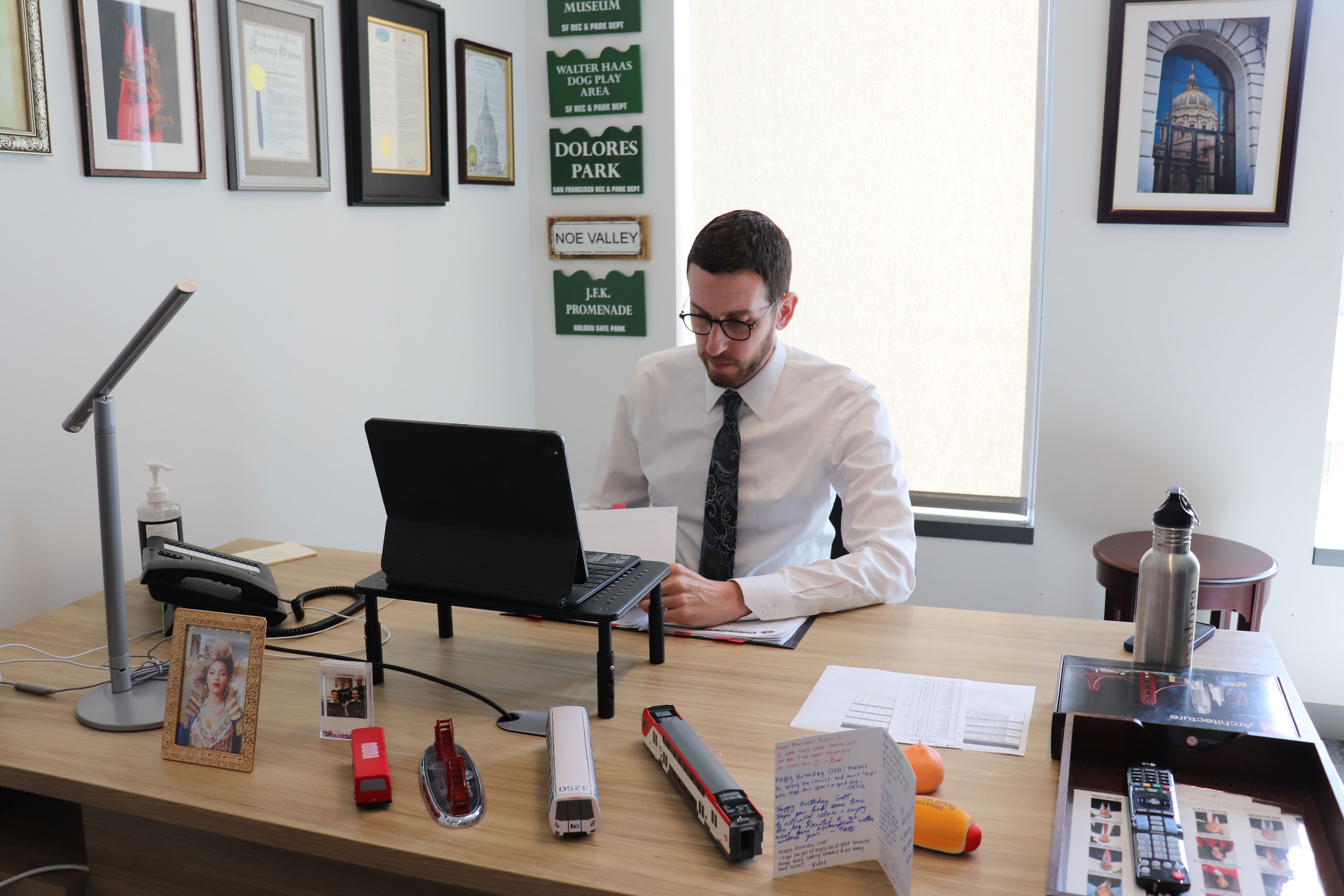 A man in a white shirt and dark tie sits behind a desk.