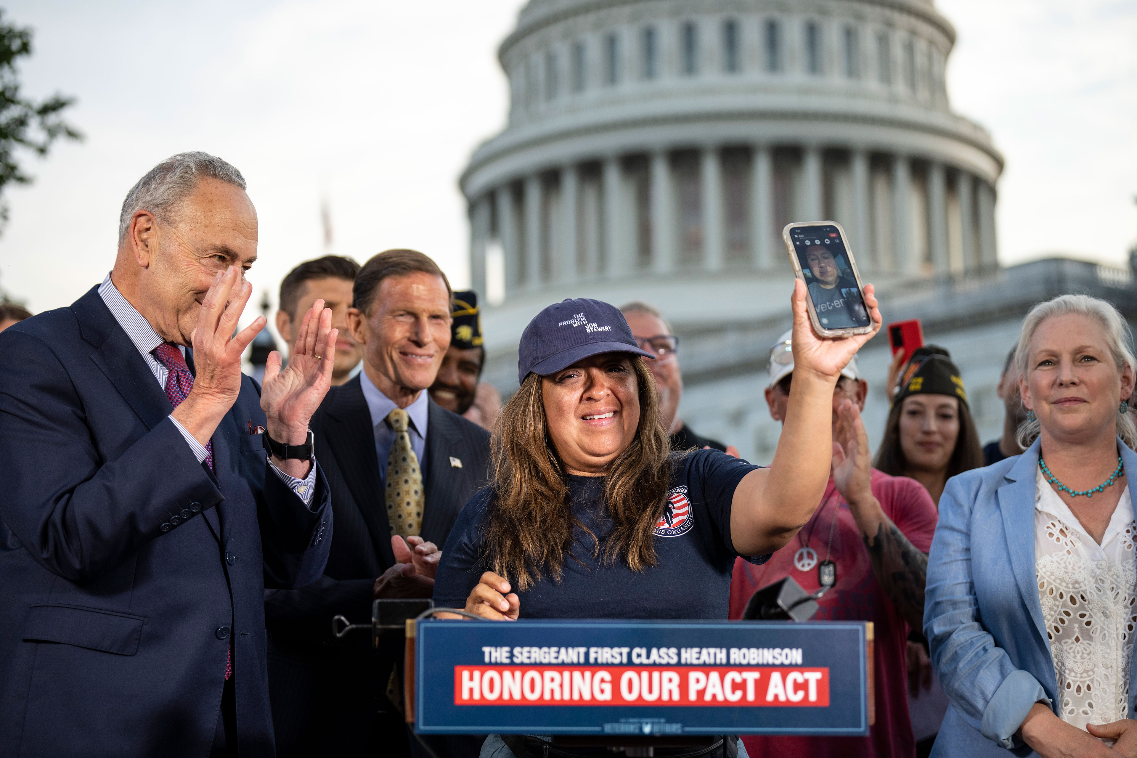 A woman in a baseball cap stands at a lecture in front of the Capitol building, surrounded by people, and holds up a phone with a man's face visible on it.