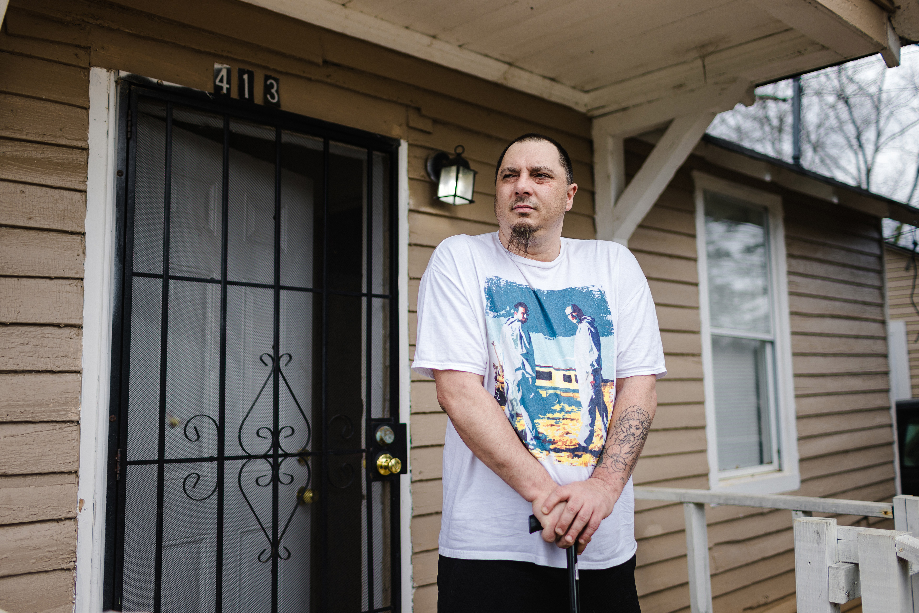 A photo shows a man standing for a portrait outside his home. He is holding a cane.