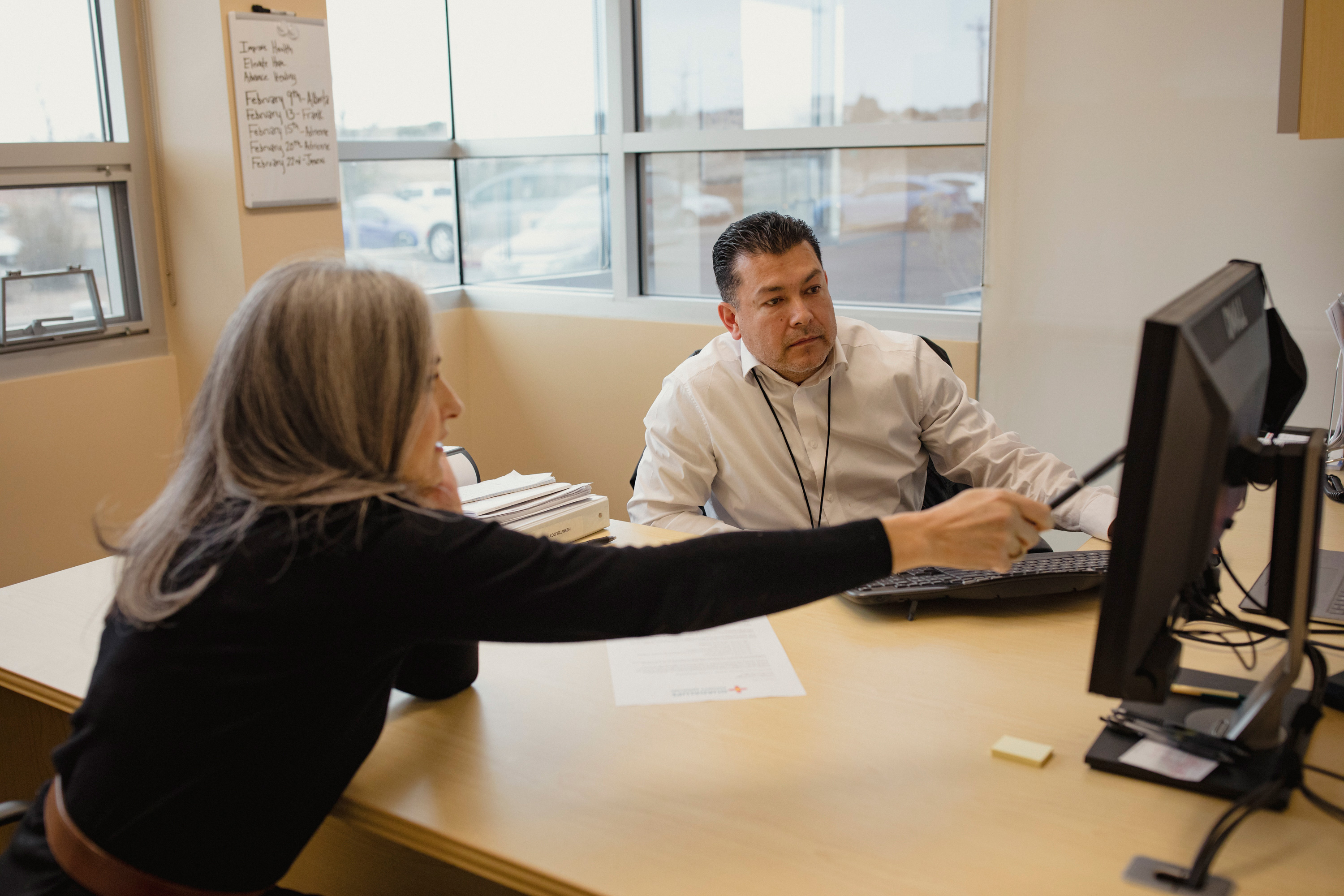 A photo shows a woman pointing with a pen to a computer monitor as a man sitting across from her listens.