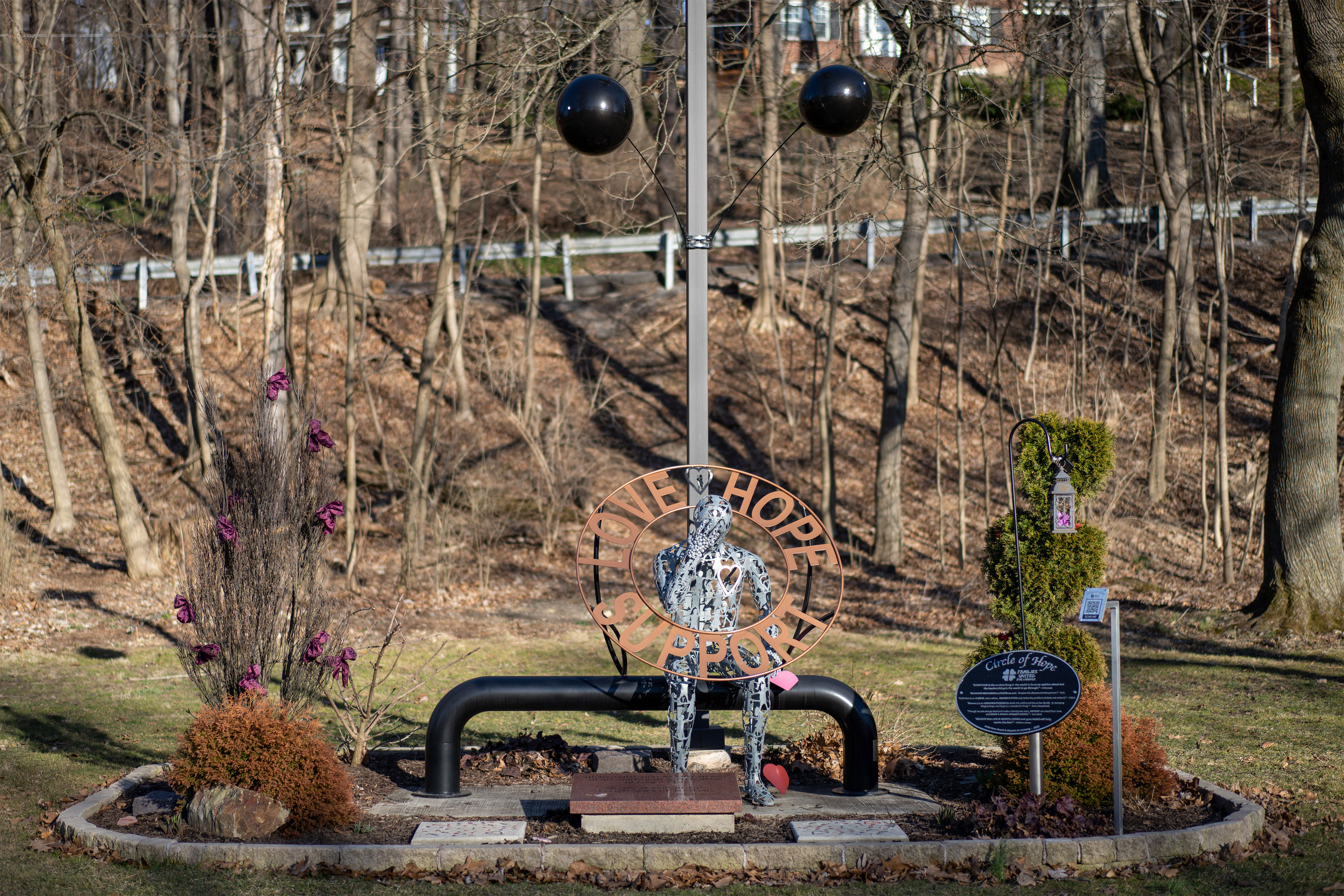 A metal statue of a person bending forward, head on hand, is encircled by a copper circle that reads, Love Hope Support. The statue is in a park surrounded by bare trees, grass, and shrubs.