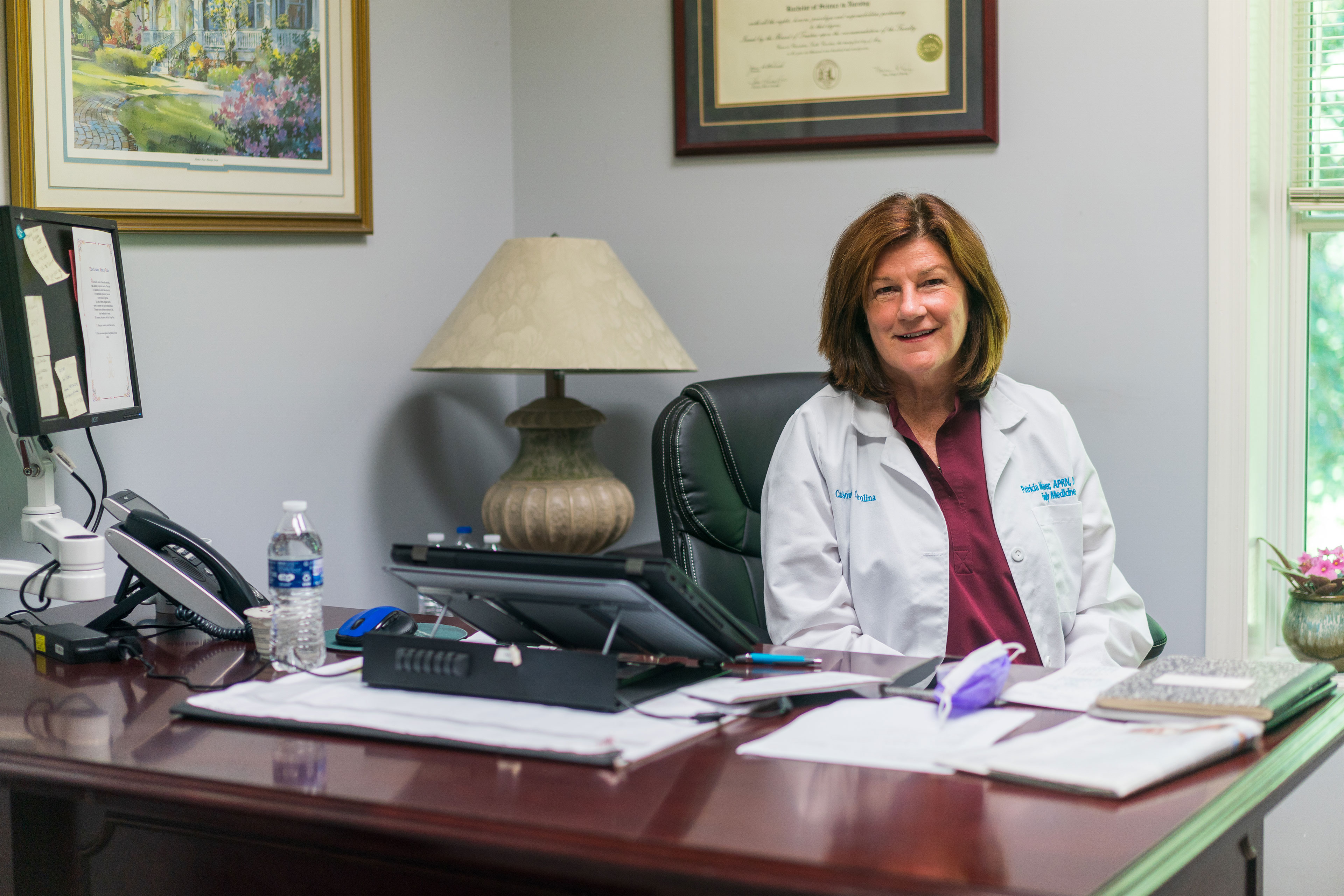 A woman in a maroon shirt and white lab coat sits behind a desk in an office and looks at the camera.