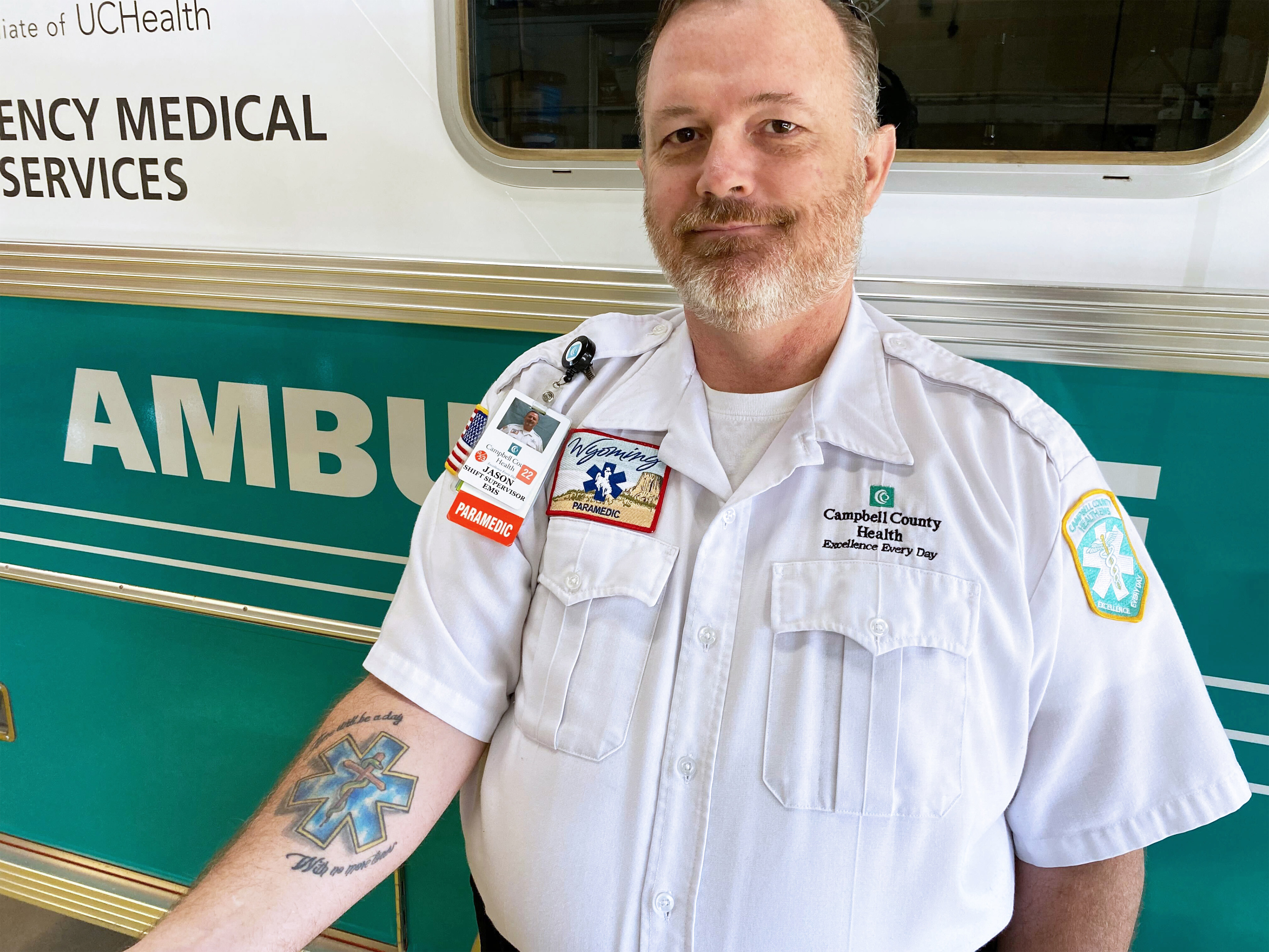 A photo of a male paramedic showing an EMS tattoo on his forearm in front of an ambulance.