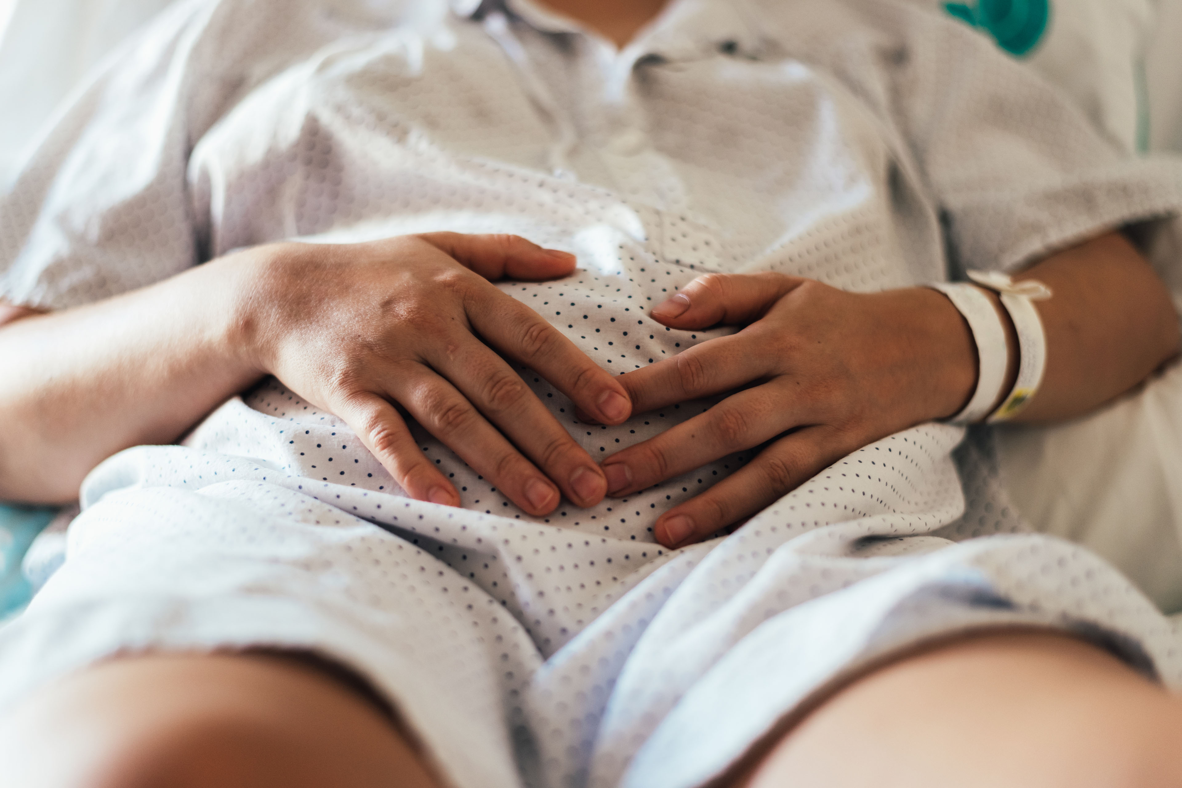 A woman in a hospital bed and gown is holding her stomach. The photo is cropped so only her stomach and hands are visible.