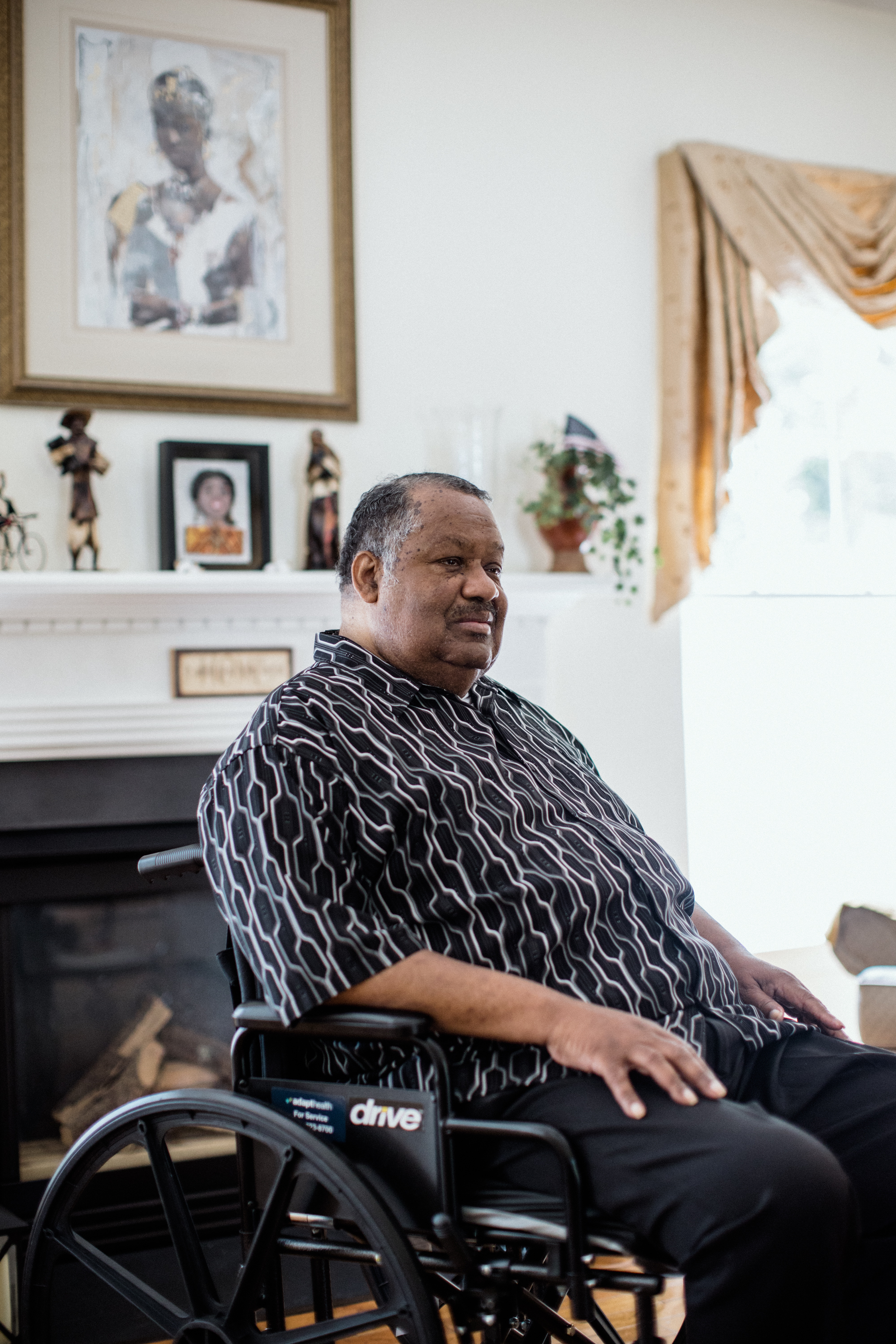 Thomas Greene is sitting in a wheelchair in his home, which is brightly lit by sunlight. He looks away from the camera.