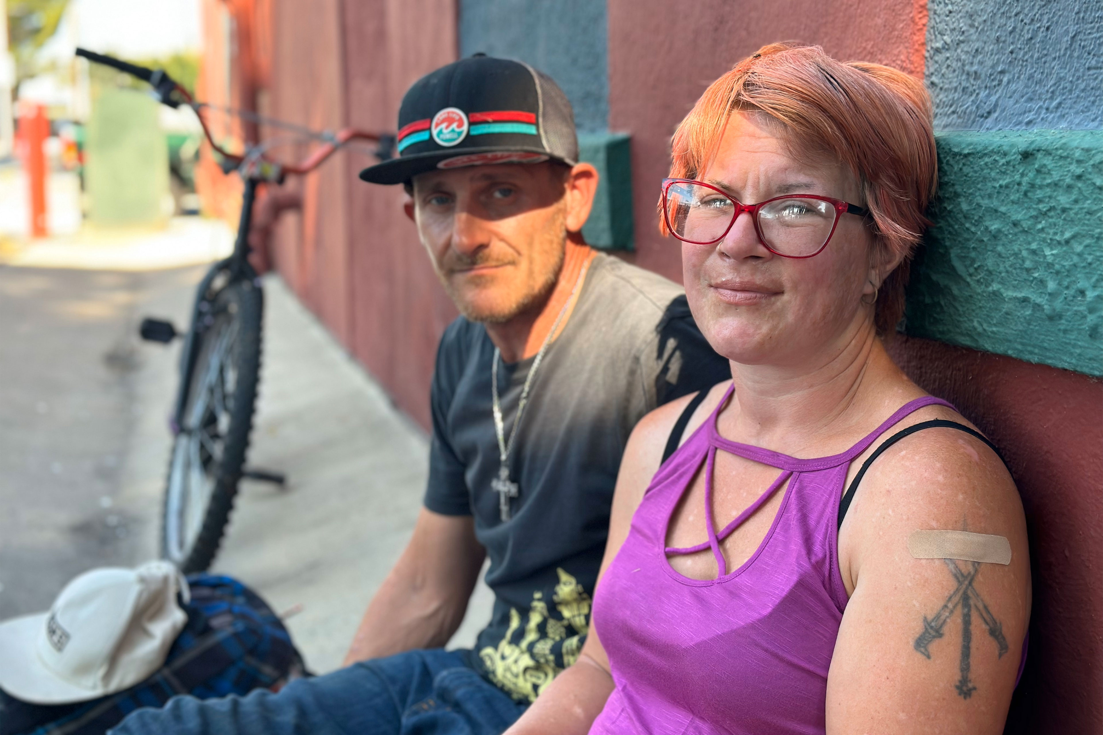 A woman with short hair wearing red glasses and a purple tank top sits beside a man wearing a baseball cap and a cross around his neck. They sit on a sidewalk and lean against a building while looking at the camera.