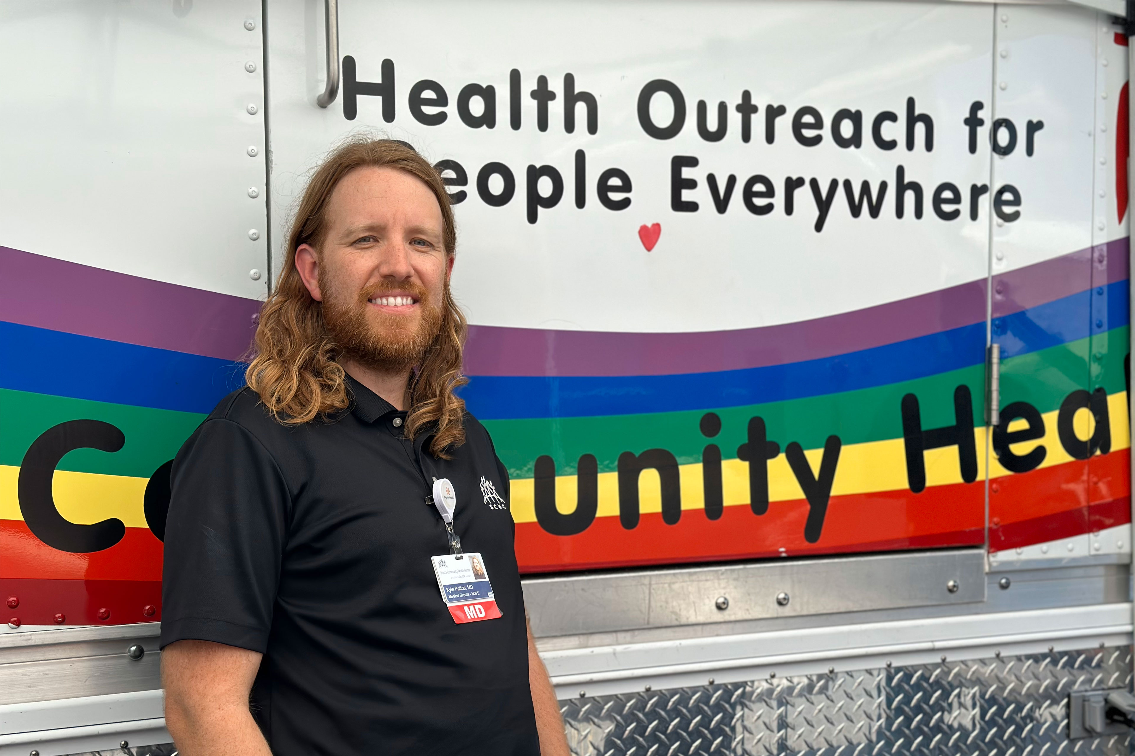 A man with shoulder-length hair wearing a black polo shirt and identification badge stands in front of a van and looks at the camera. The van is printed with the words, "Health Outreach for People Everywhere."