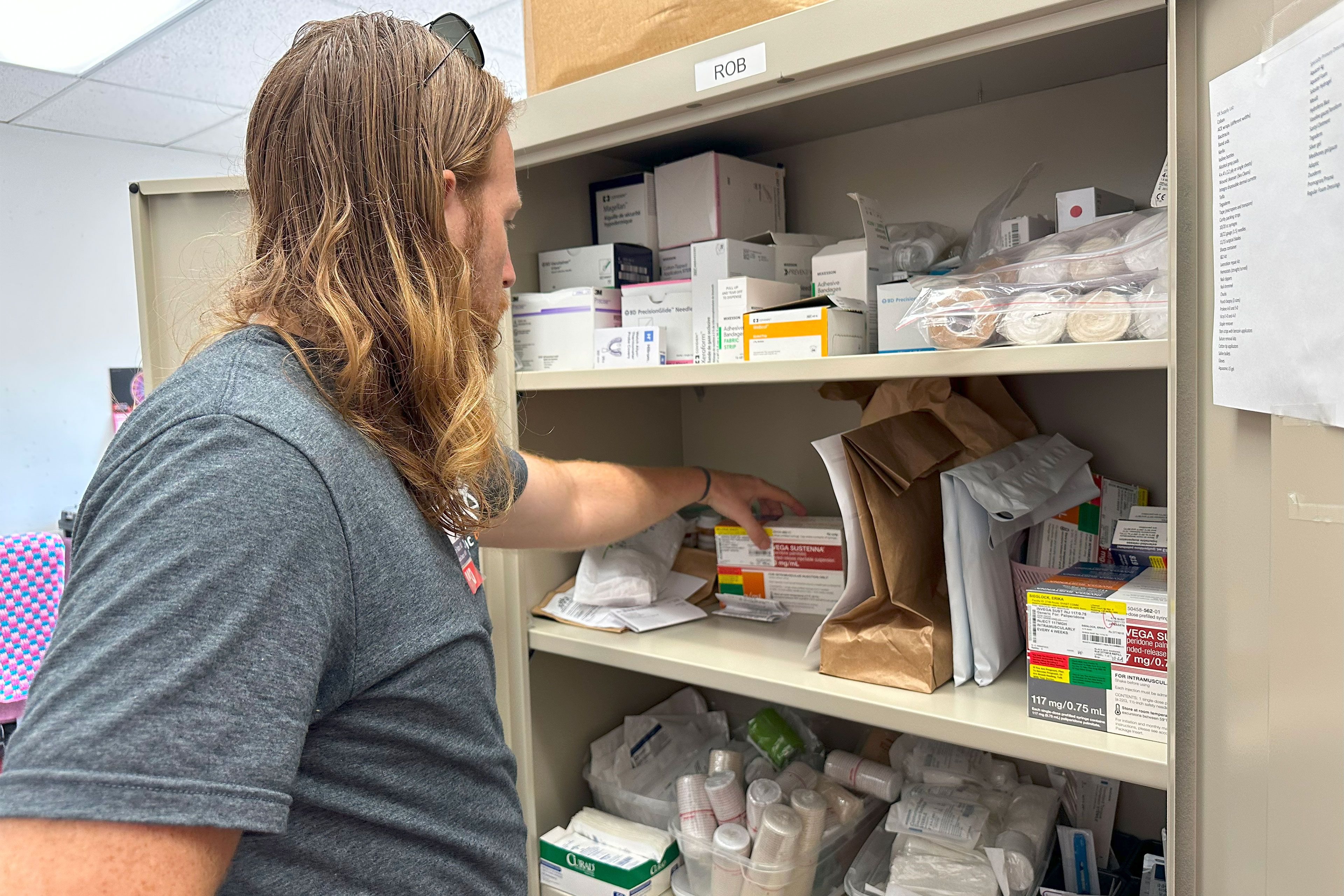 A man with shoulder-length hair below his shoulders reaches into a metal cabinet to pick up a box.