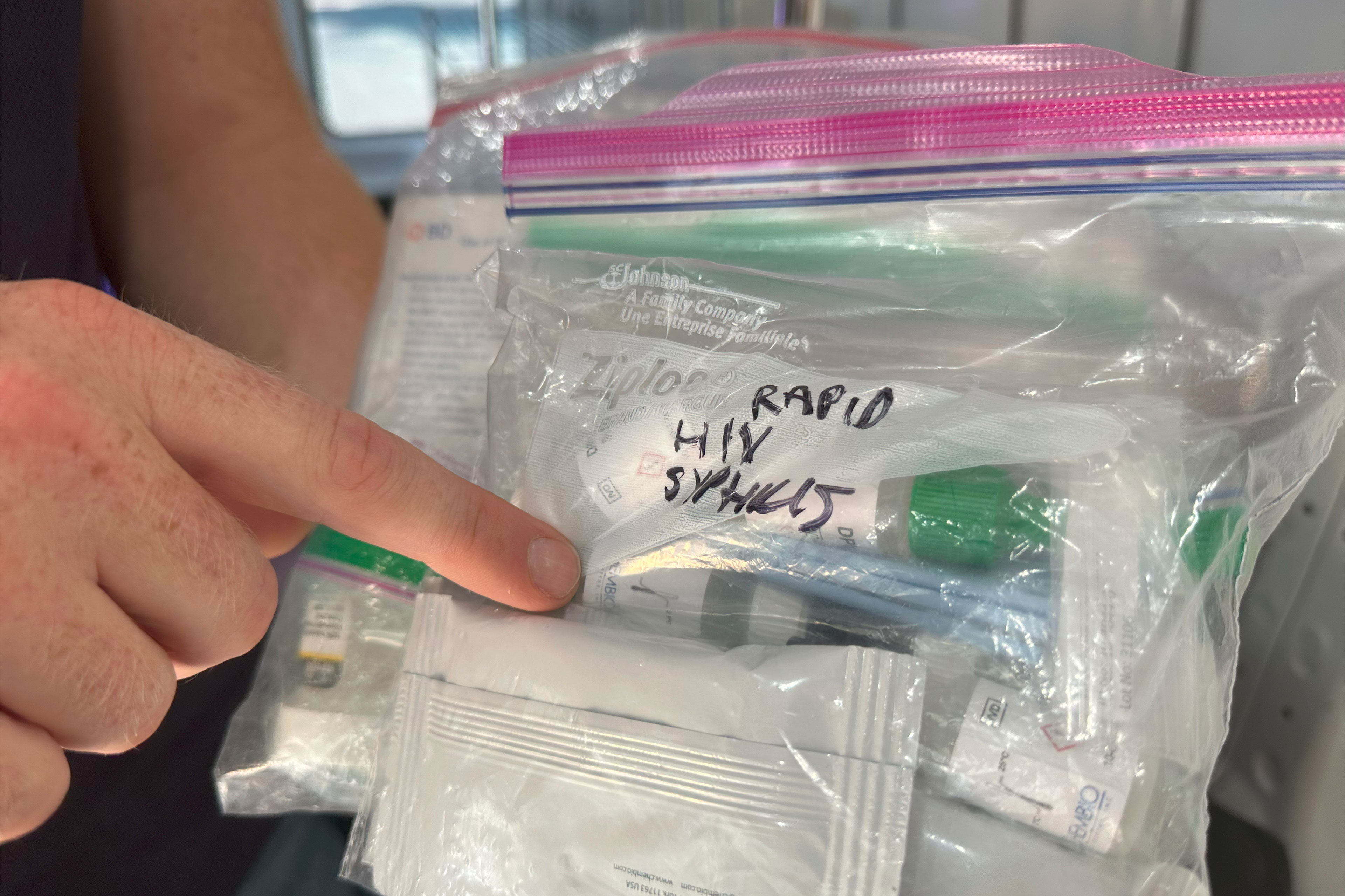 A hand points to a plastic bag containing medical supplies. Someone has written "RAPID HIV SYPHILIS" in Sharpie on the bag.