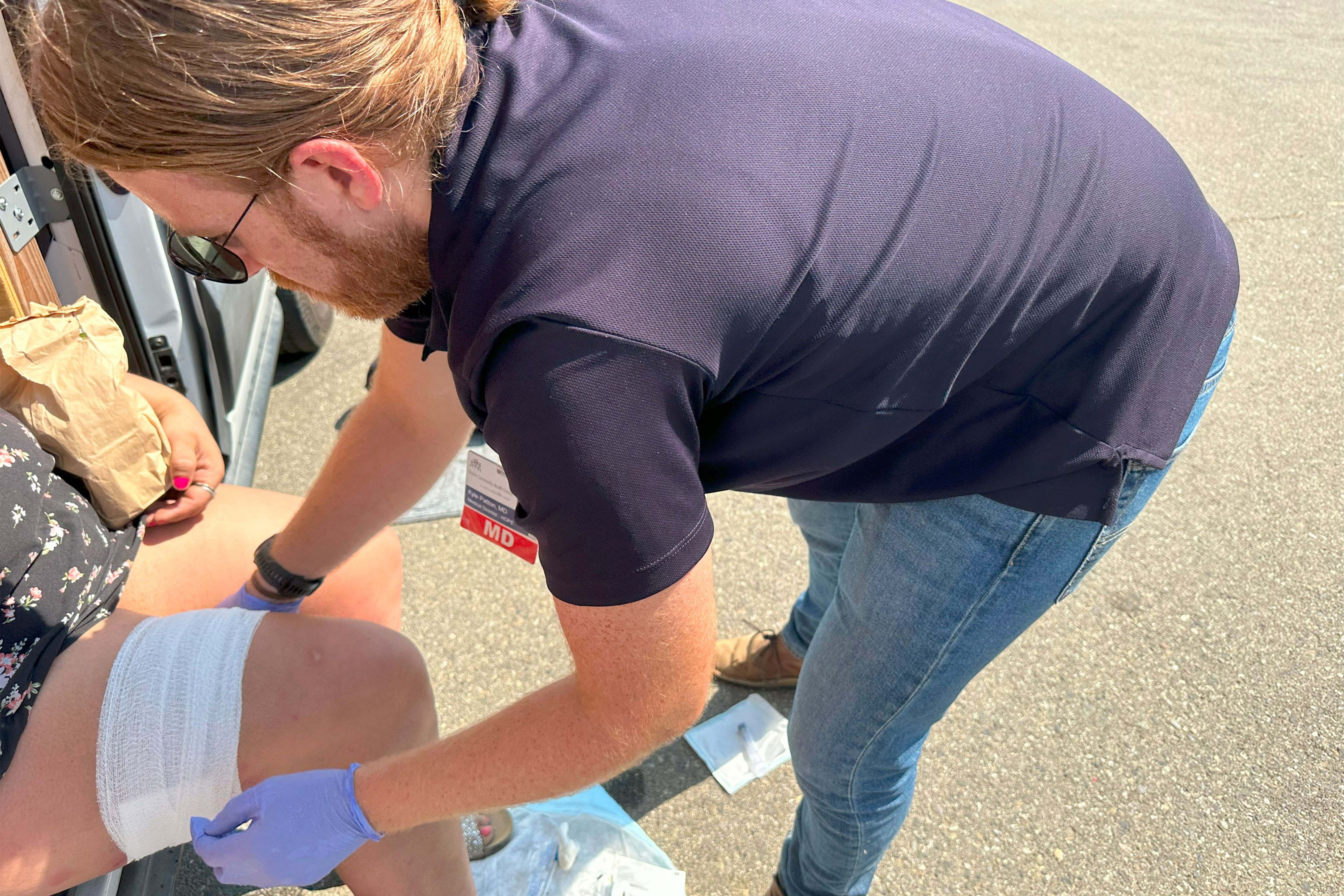 A man leans over to adjust the bandage on an unidentifiable woman's thigh as she sits on the edge of a van.
