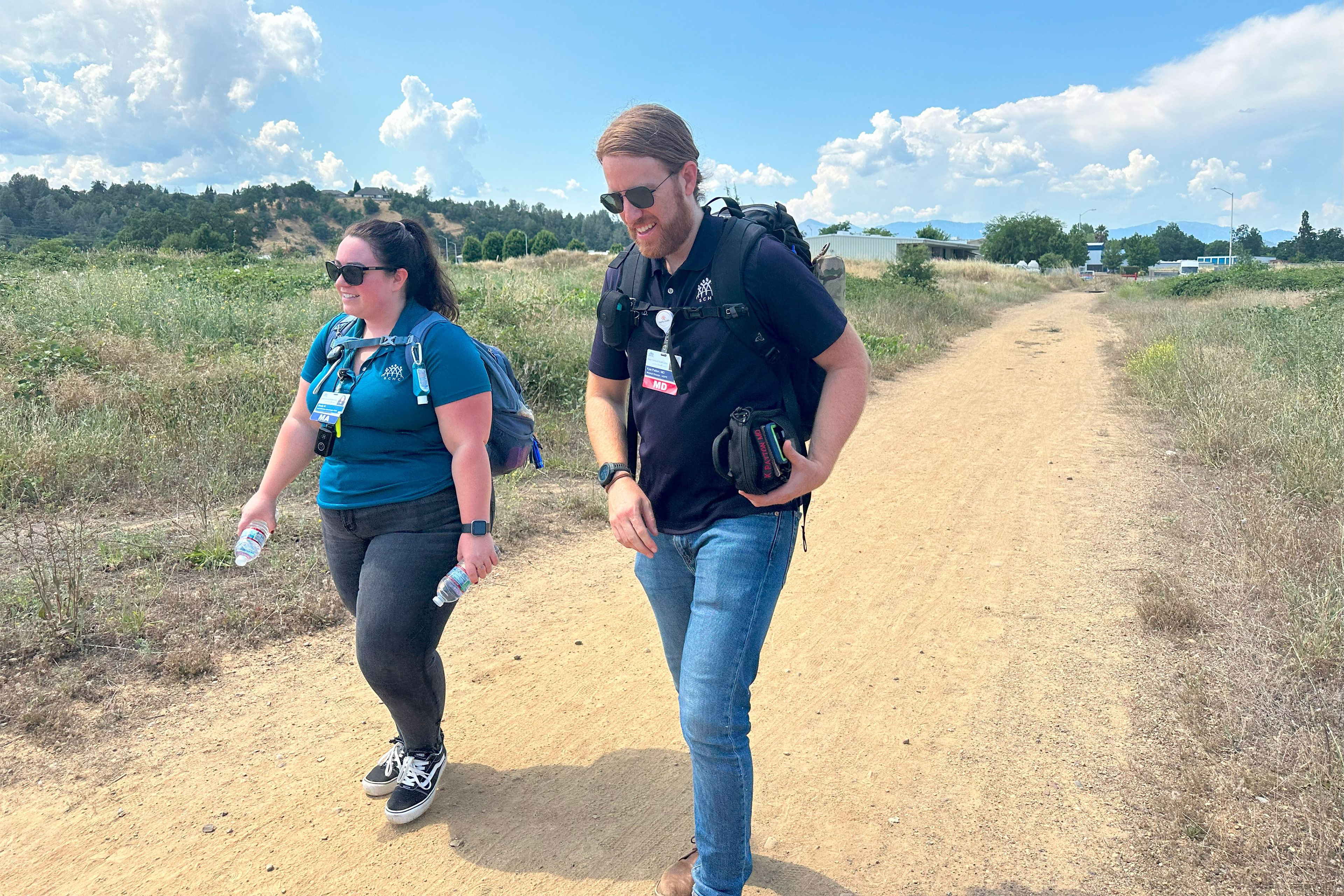 A man and a woman — both wearing sunglasses, identification badges, and backpacks — walk along a dirt path through a meadow on a sunny day.