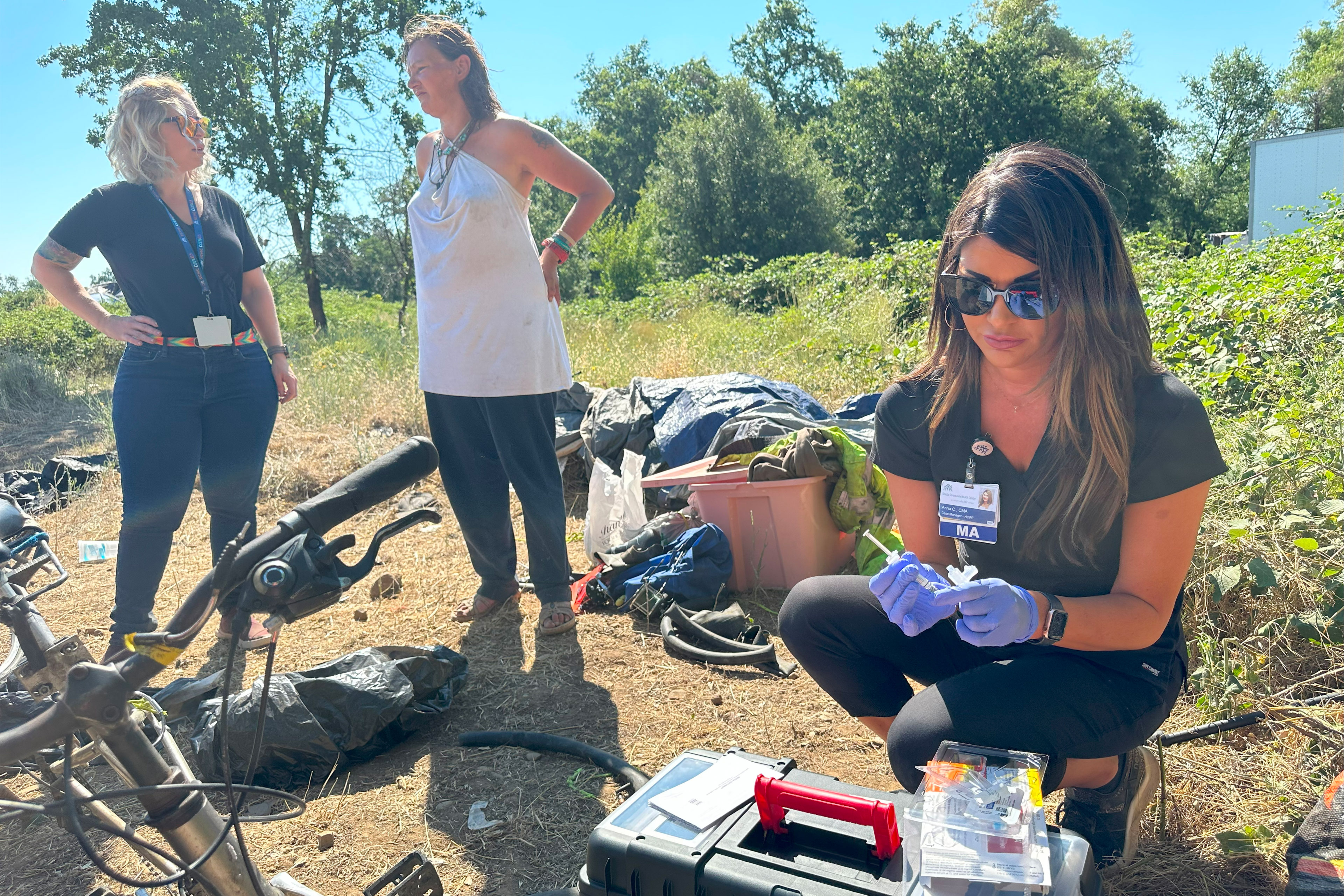 A woman wearing sunglasses and black scrubs with a name badge attached to her top squats beside a plastic box and prepares a syringe. Two women a short distance behind have a conversation.