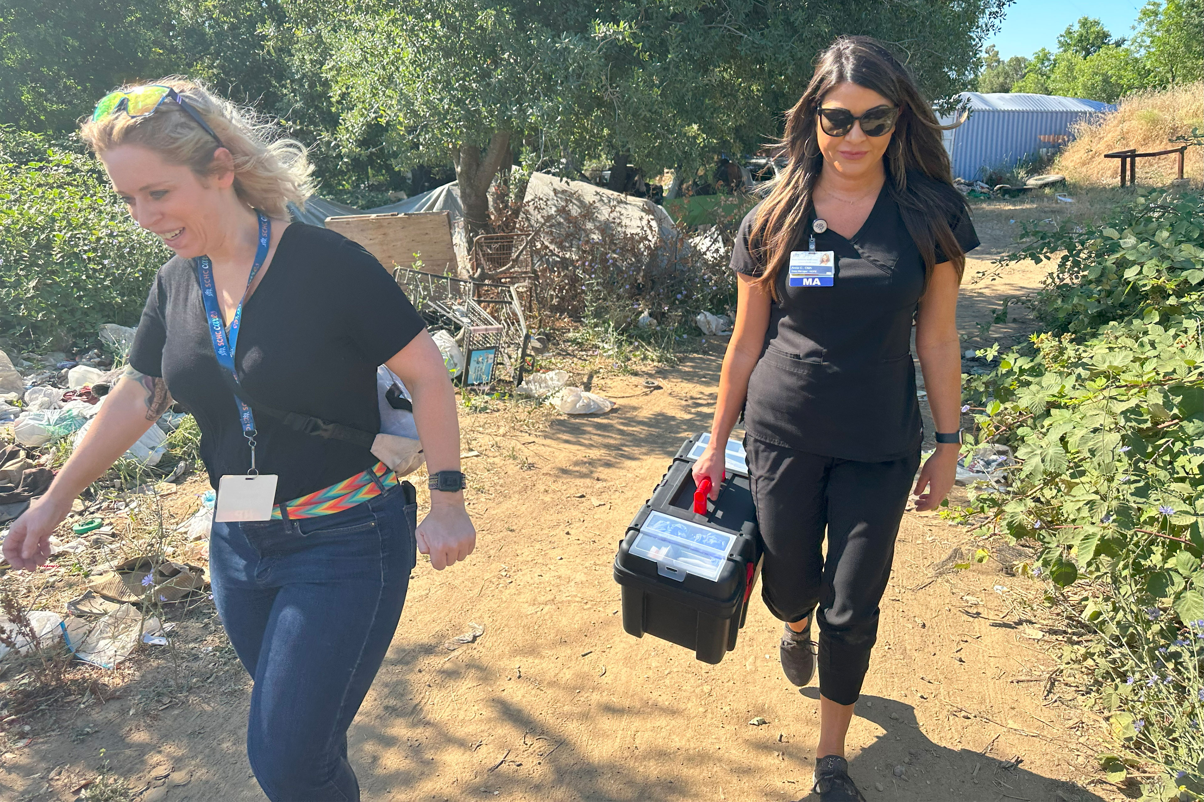 Two women walk along a path with a tent, sideways shopping cart, and trash behind them. The woman on the left has blonde hair and sunglasses on top of her head. She wears a black tee-shirt, jeans with a colorful belt, and a badge on a lanyard around her neck. The woman on the right has long hard hair and carries a plastic box by the handle. She wears sunglasses and black scrubs with an ID badge attached to her top.