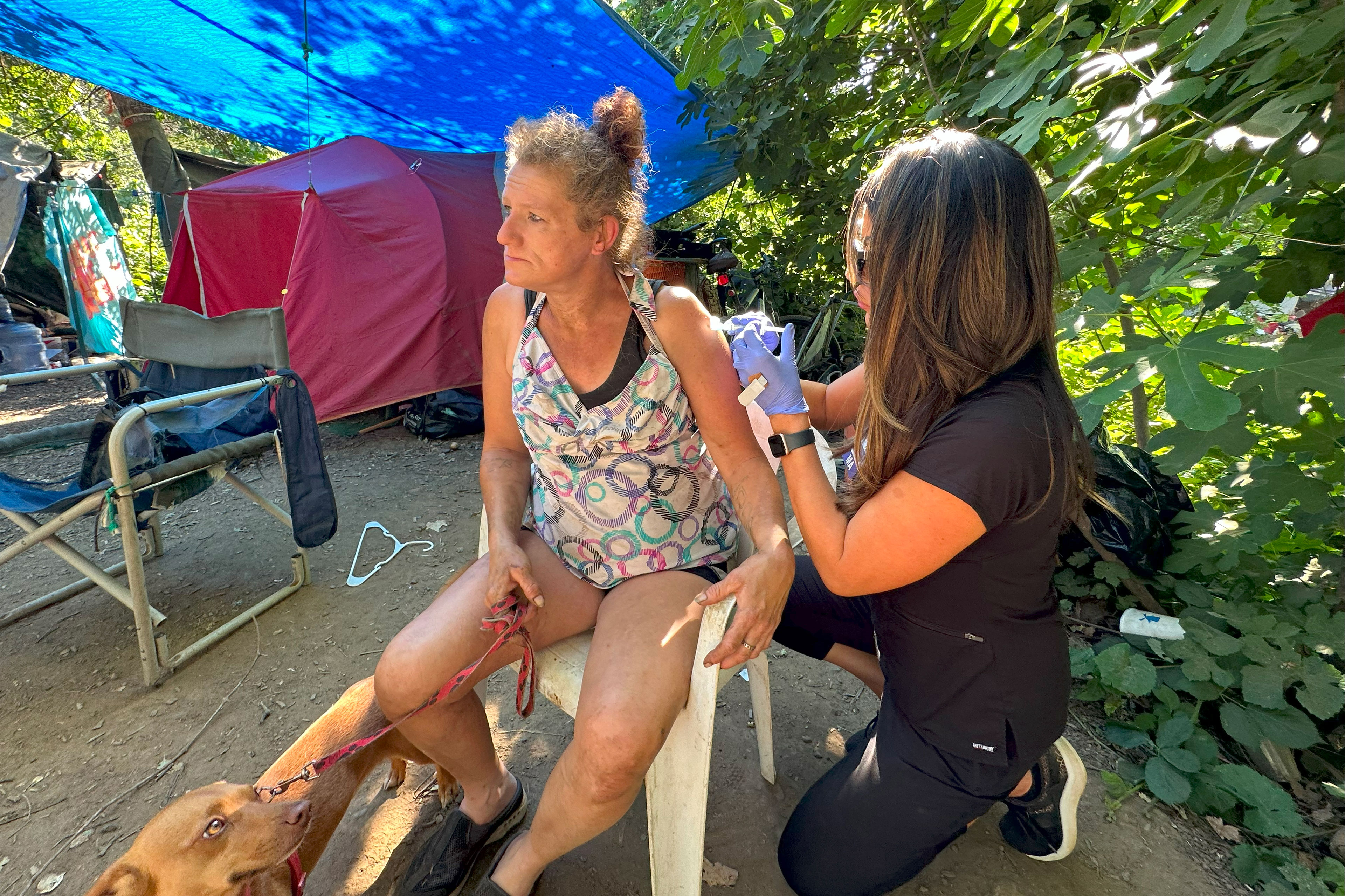 A woman in black scrubs kneels on the ground as she administers an injection to another woman sitting in a plastic chair. The seated woman holds a red leash attached to a small brown dog and looks away from the shot.