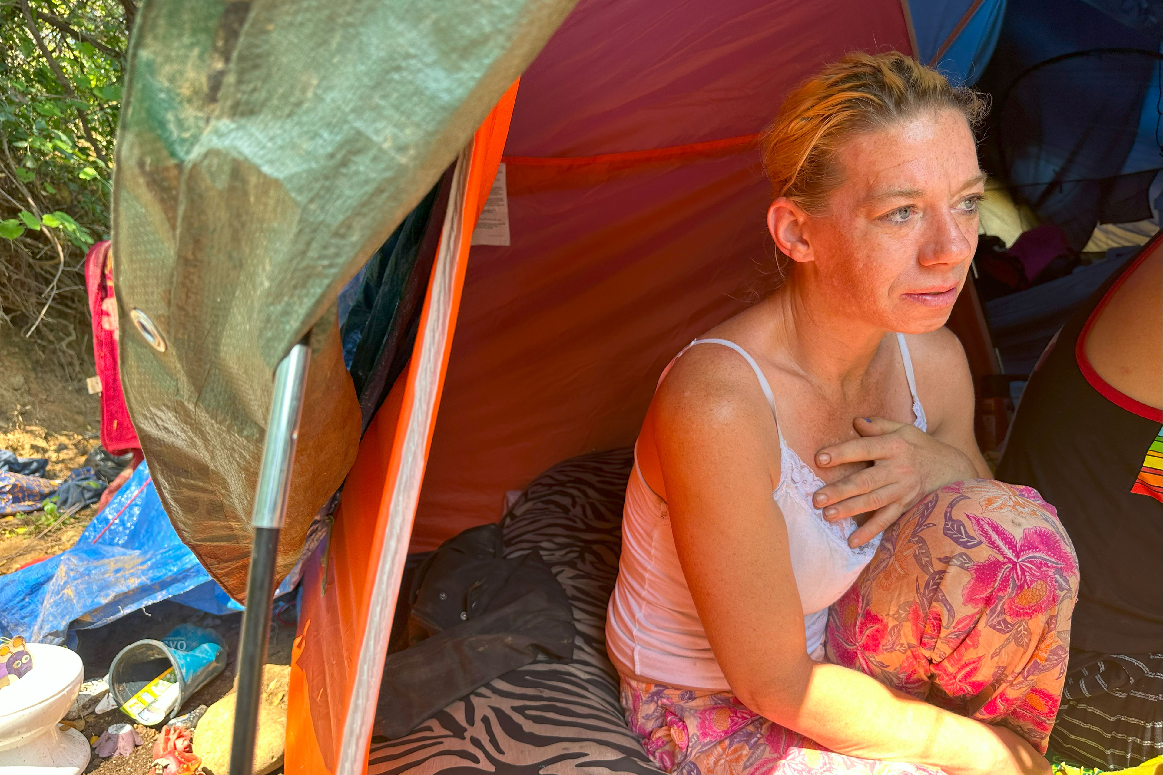 A woman sits in a tent and and looks at something off-camera. She wears a white tank top and pants with a floral pattern. She holds one hand to her chest.
