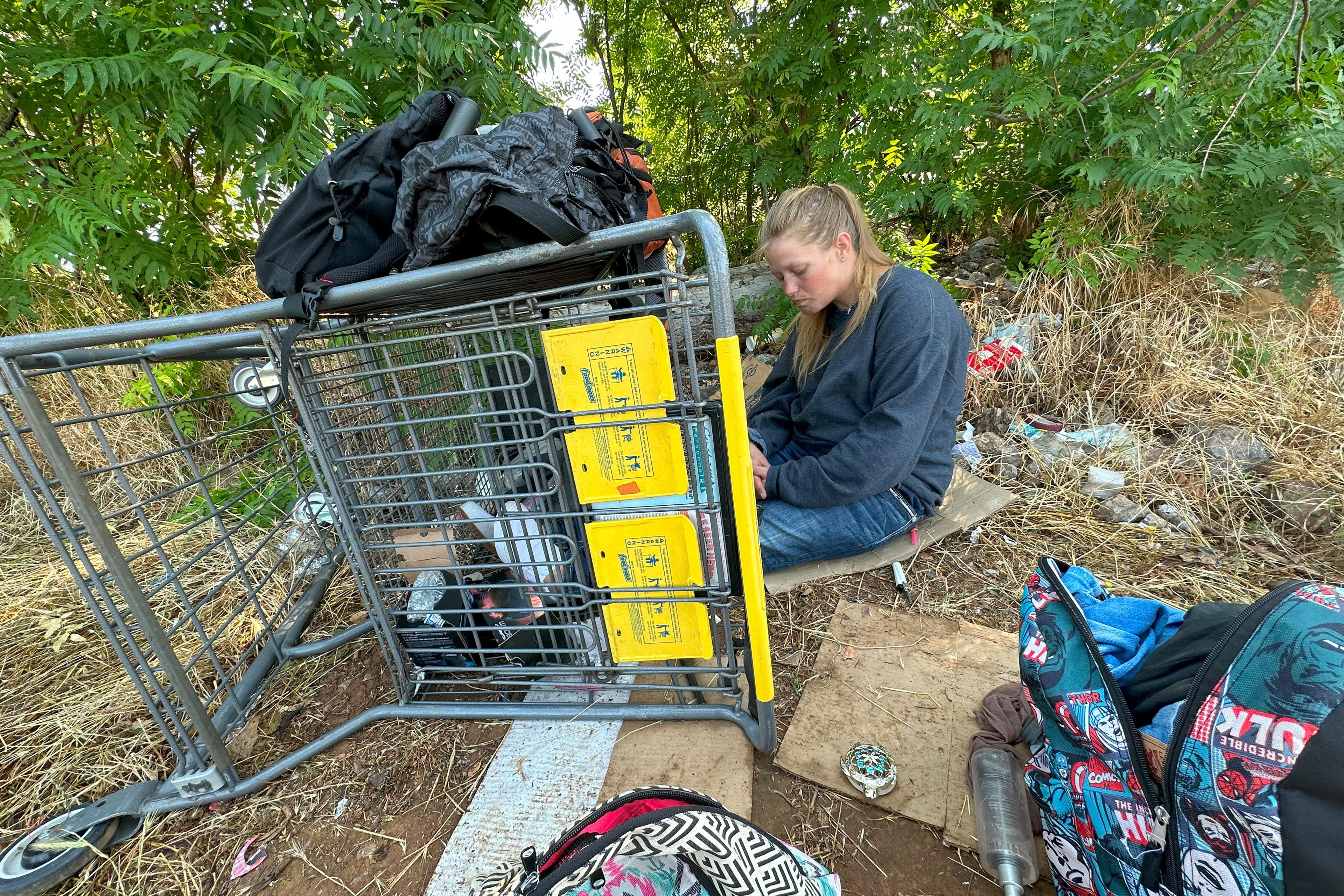 A woman with long blonde hair and wearing jeans and a dark-colored sweatshirt sits on a piece of cardboard outside. Her outstretched legs are in a shopping cart on its side on the ground. There are bags, other pieces of cardboard, and trash around the woman and in the cart.