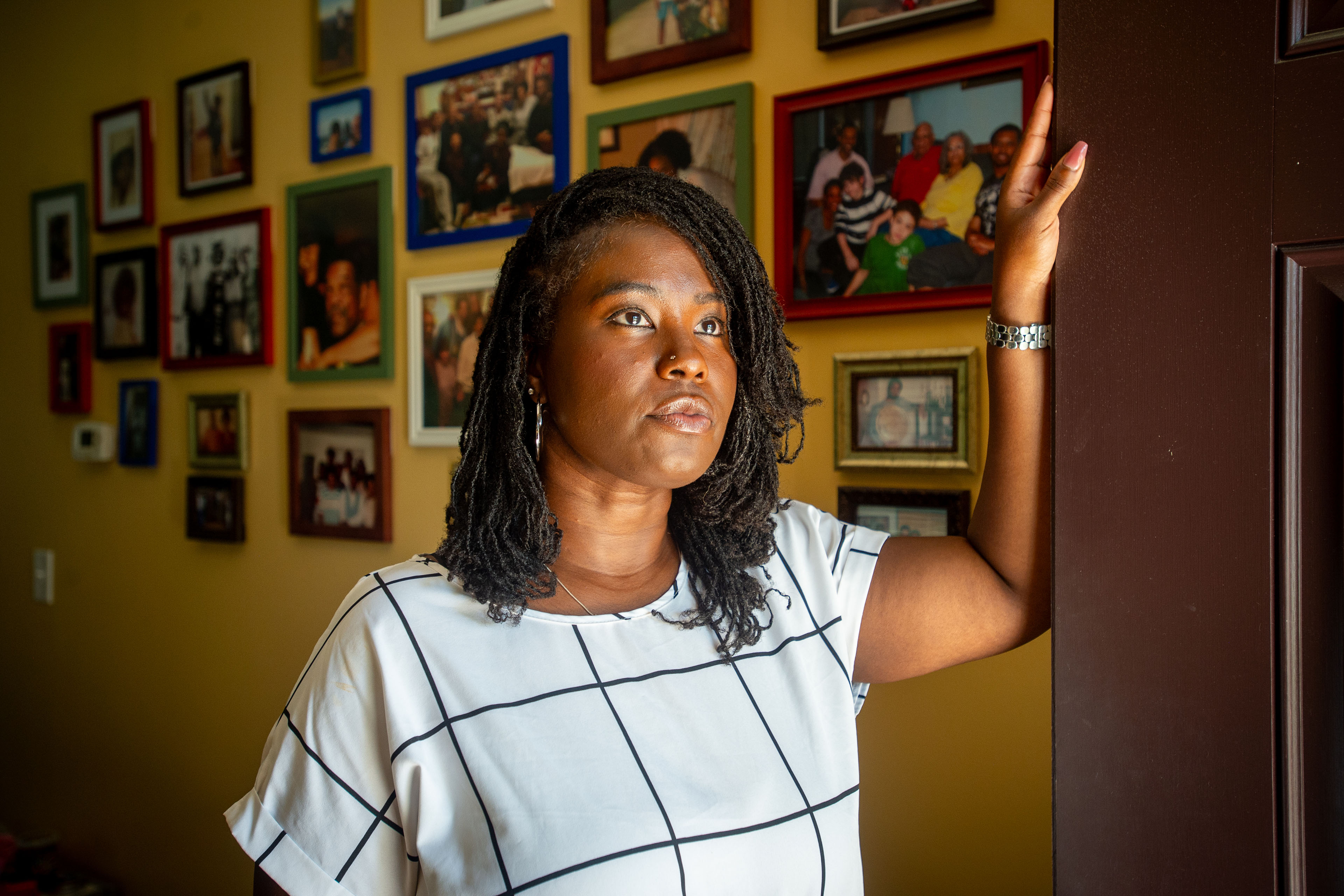Charity Watkins stands in the doorway of her home, looking outside. Framed photographs cover the wall behind her in a tiled pattern.