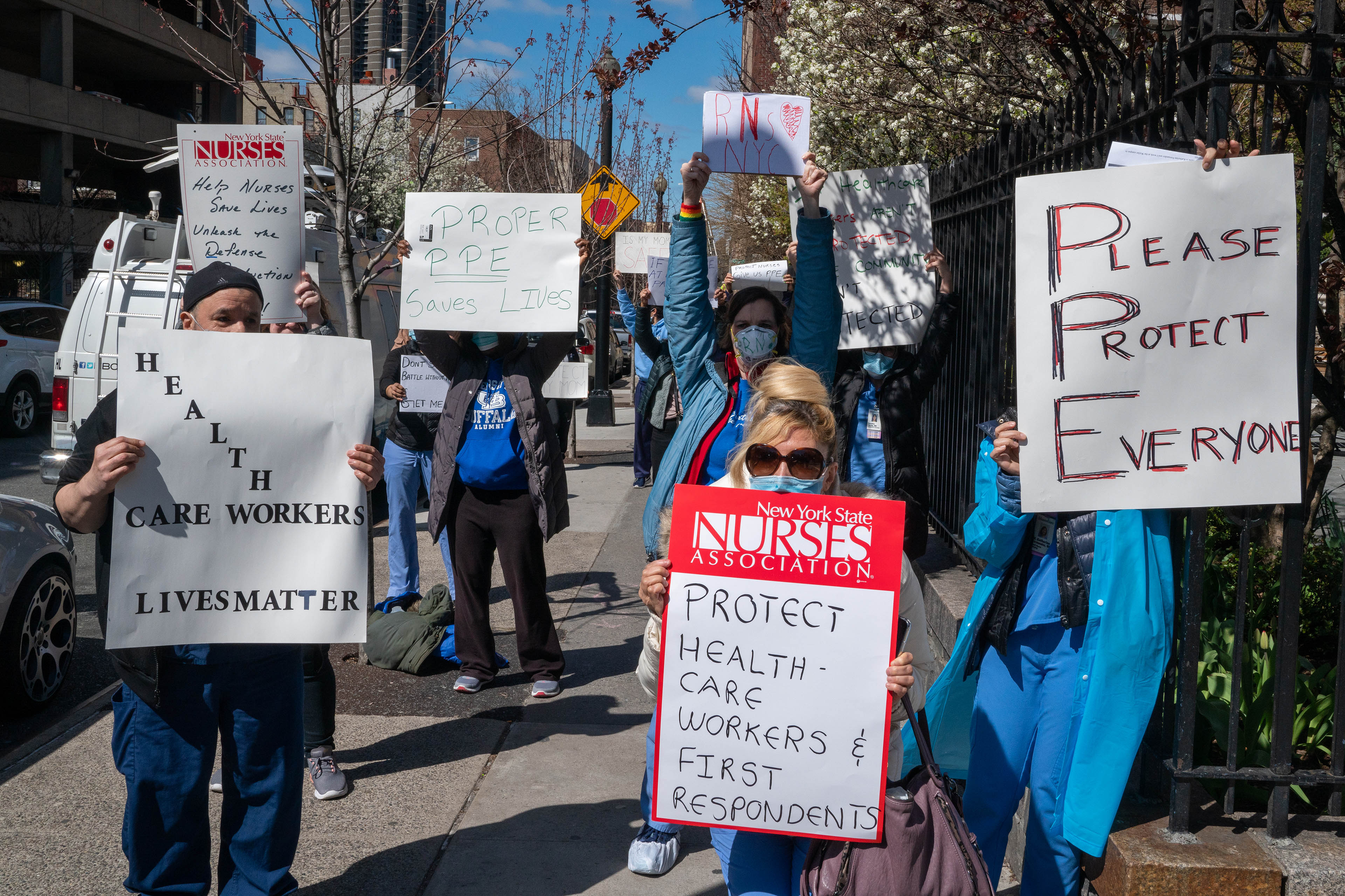 Public health workers, doctors, and nurses protest outside a New York City hospital, holding signs. They read, from left to right, "Help nurses save lives," "health care workers lives matter," "proper PPE saves lives," "protect health care workers and first respondents," and "Please Protect Everyone."
