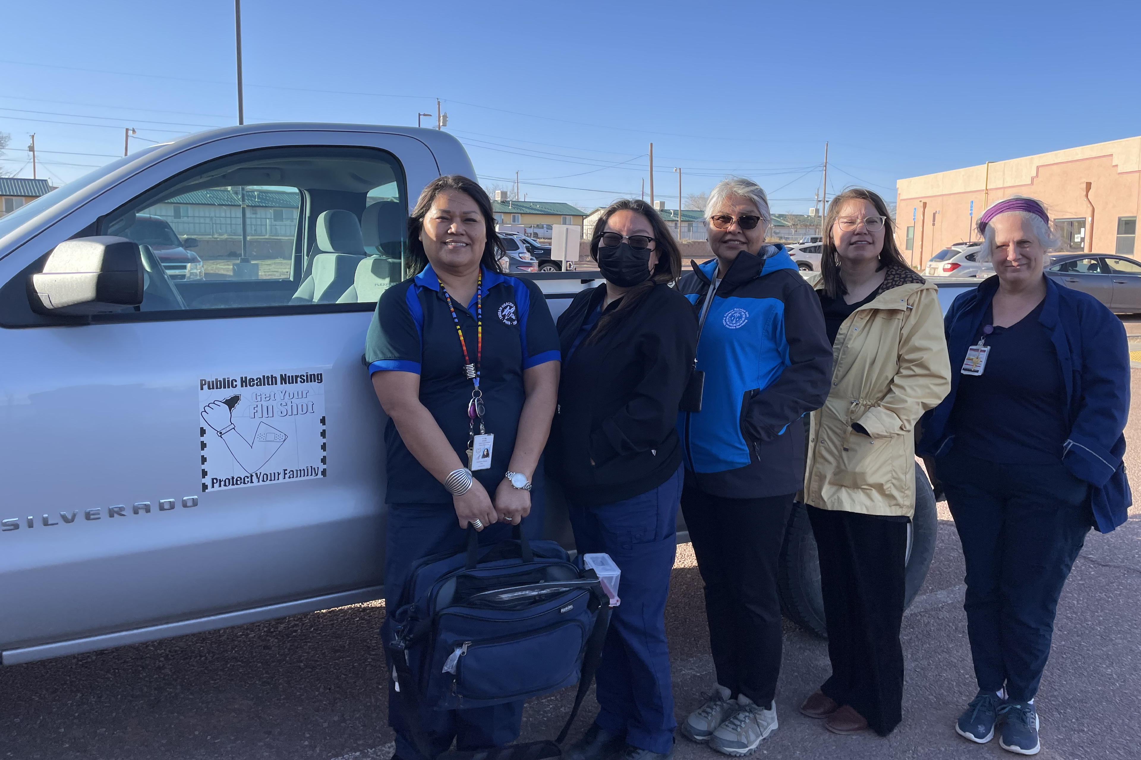 A group of people stand in front of a pick-up truck. They are wearing scrubs and jackets. One is wearing a mask.