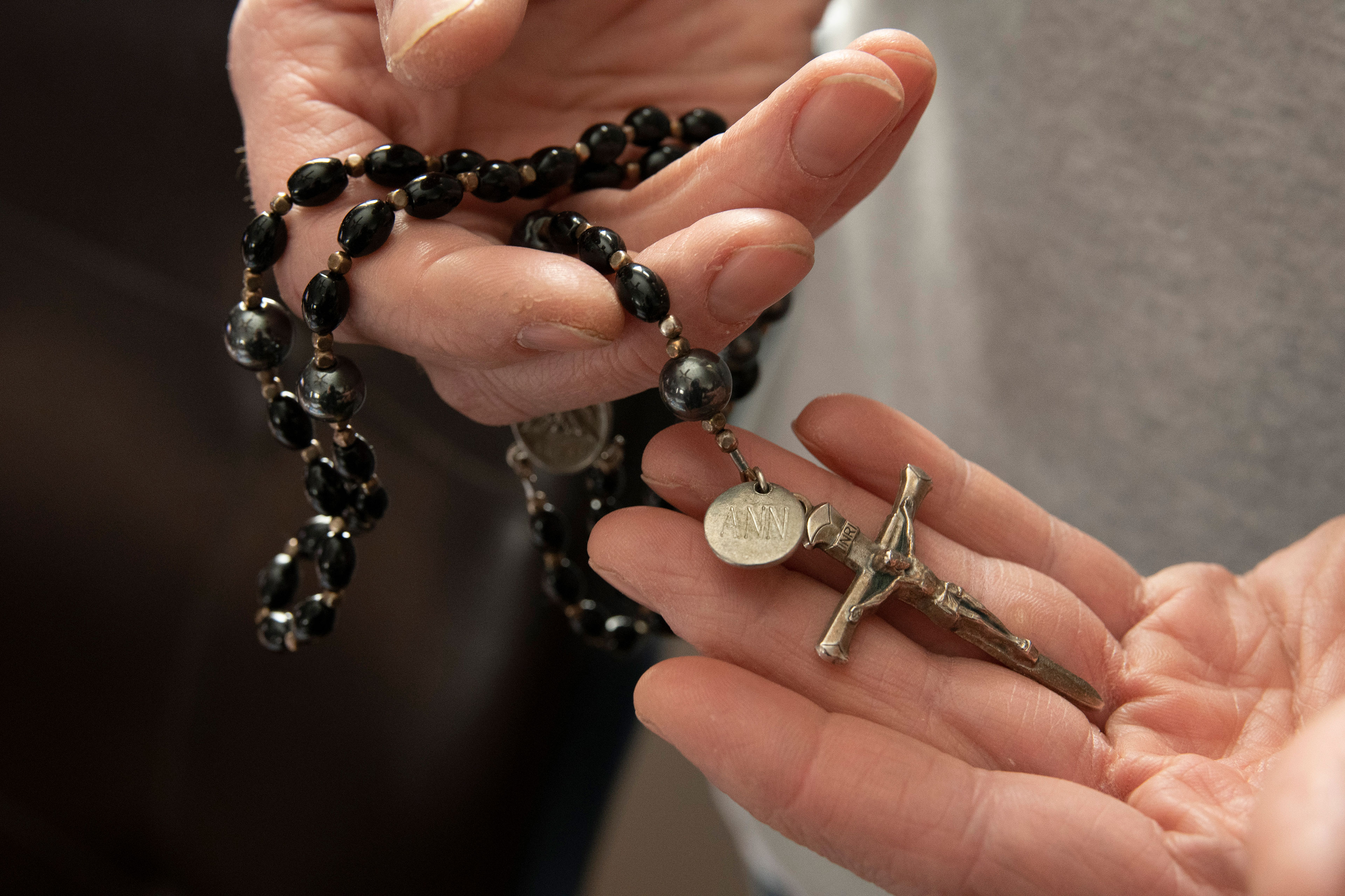 A close-up image of two hands holding a rosary with dark beads and a small disc engraved with "ANN".
