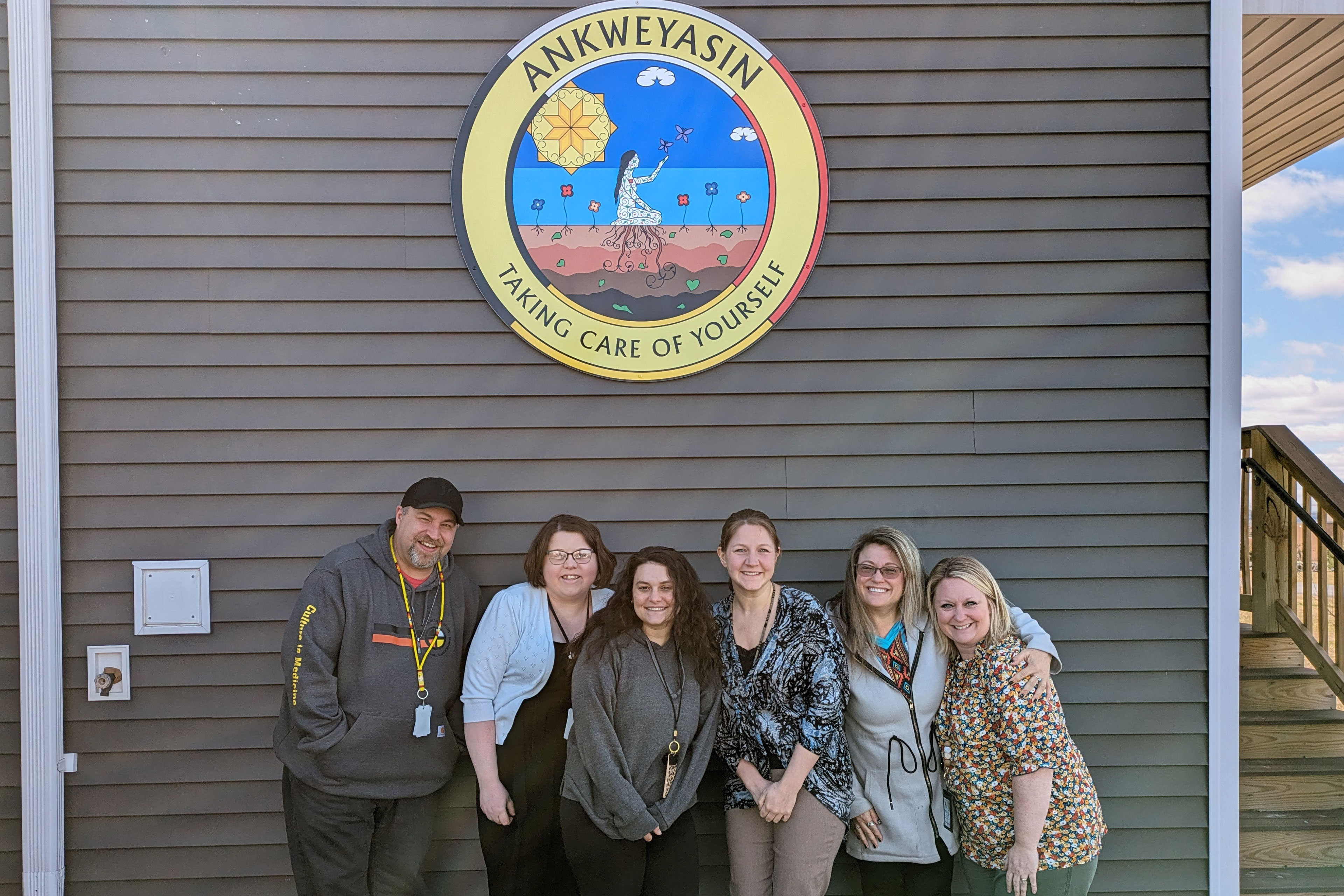 Six people stand together outside of a building, three with arms around others, and smile. Above them on the building is a round sign that reads "Ankweyasin" above an image of a person kneeling in a field of plants with the sun above, butterflies flying around their outstretched hand, and roots protruding into the ground where the person kneels. "Takaing care of yourself" reads the text at the bottom of the sign.