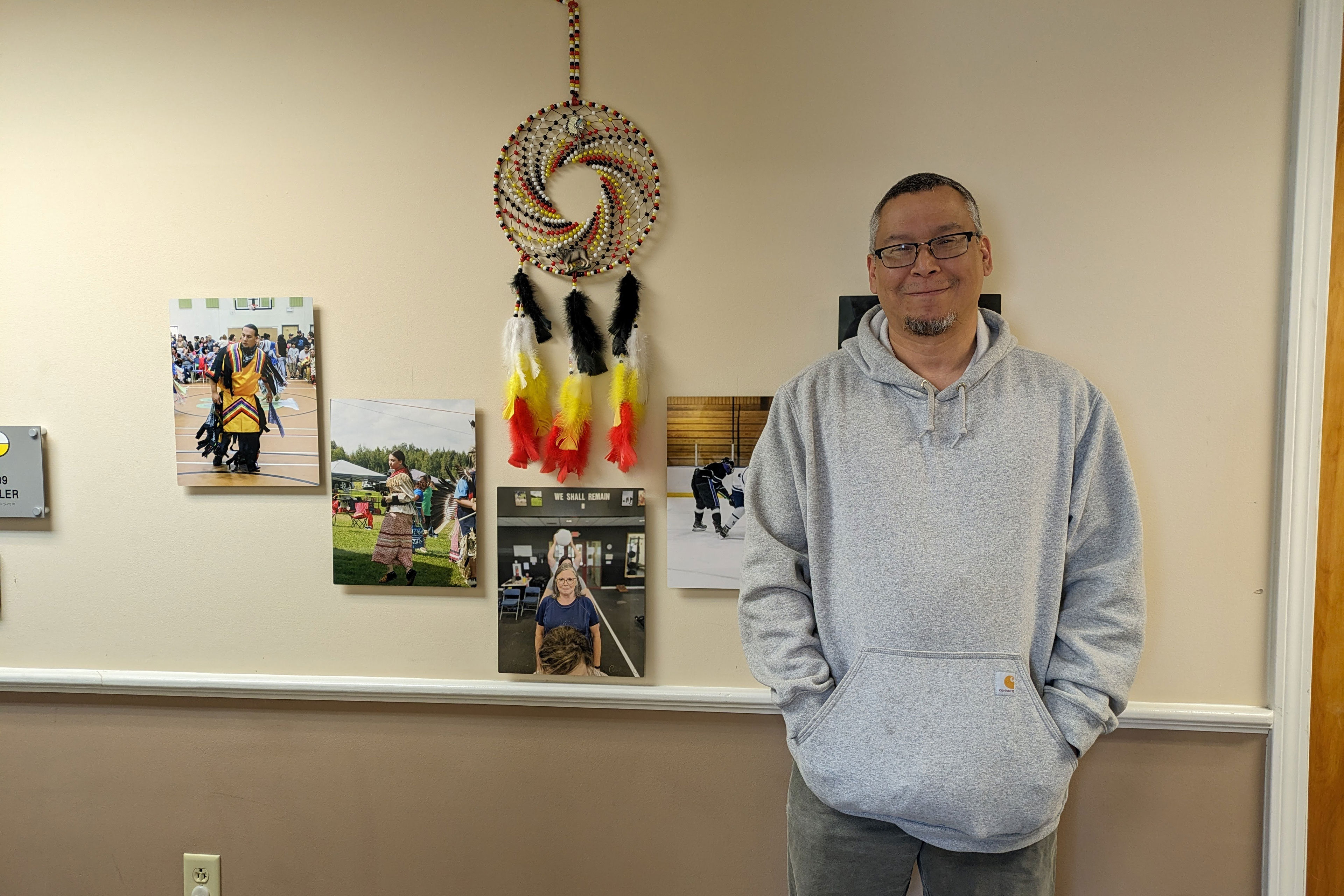 A man in a light gray hooded sweatshirt smiles at the camera while standing in front of a wall where photos and artwork are mounted.