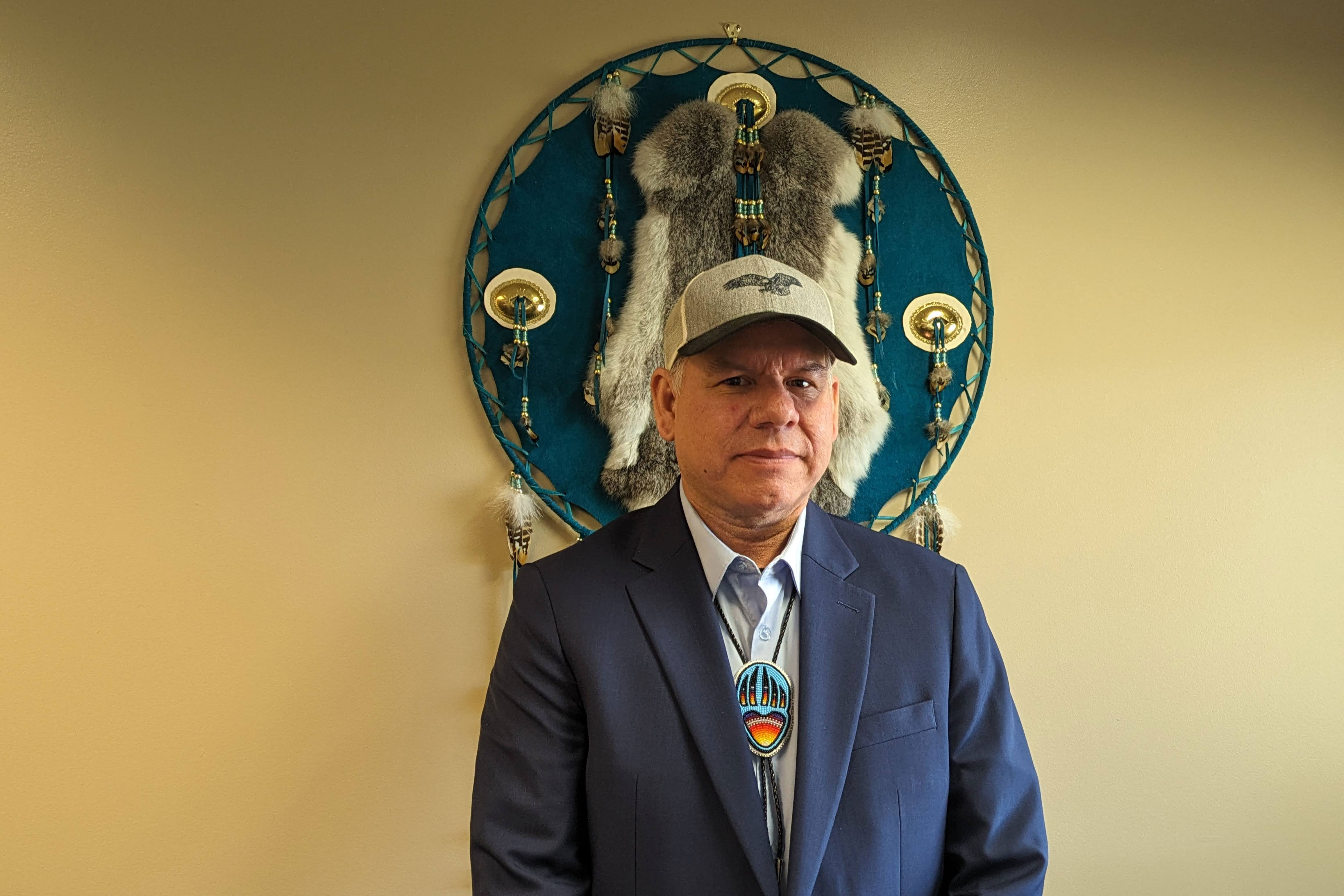 A man wearing a baseball cap, suit jacket, and beaded bolo tie stands and looks at the camera. A large work of art that includes fur and feathers is mounted on the wall behind him.