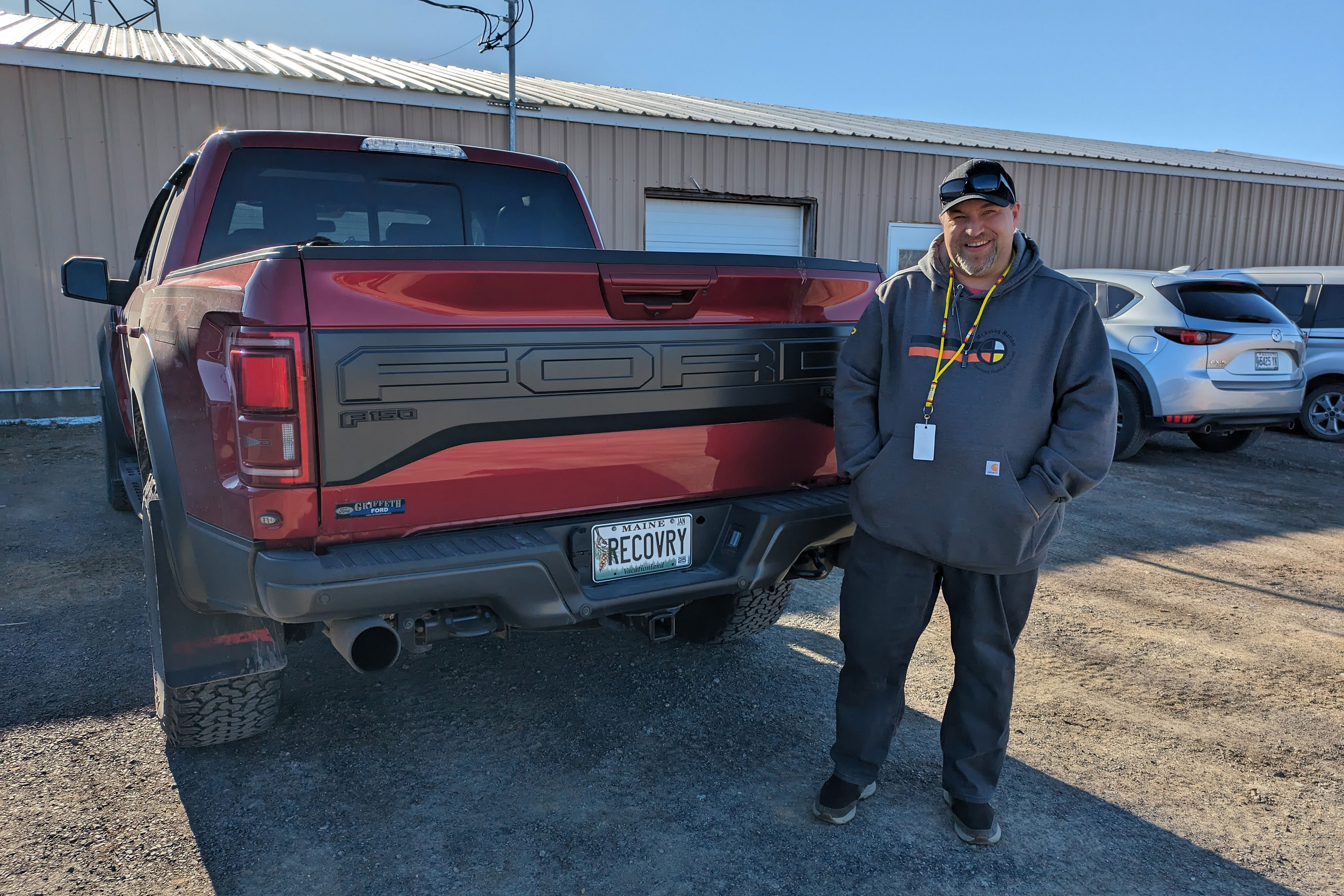 A man in a dark gray hooded sweatshirt and baseball cap stands at the back of a parked dark red Ford pickup truck. The Maine license plate on the truck reads "RECOVRY".