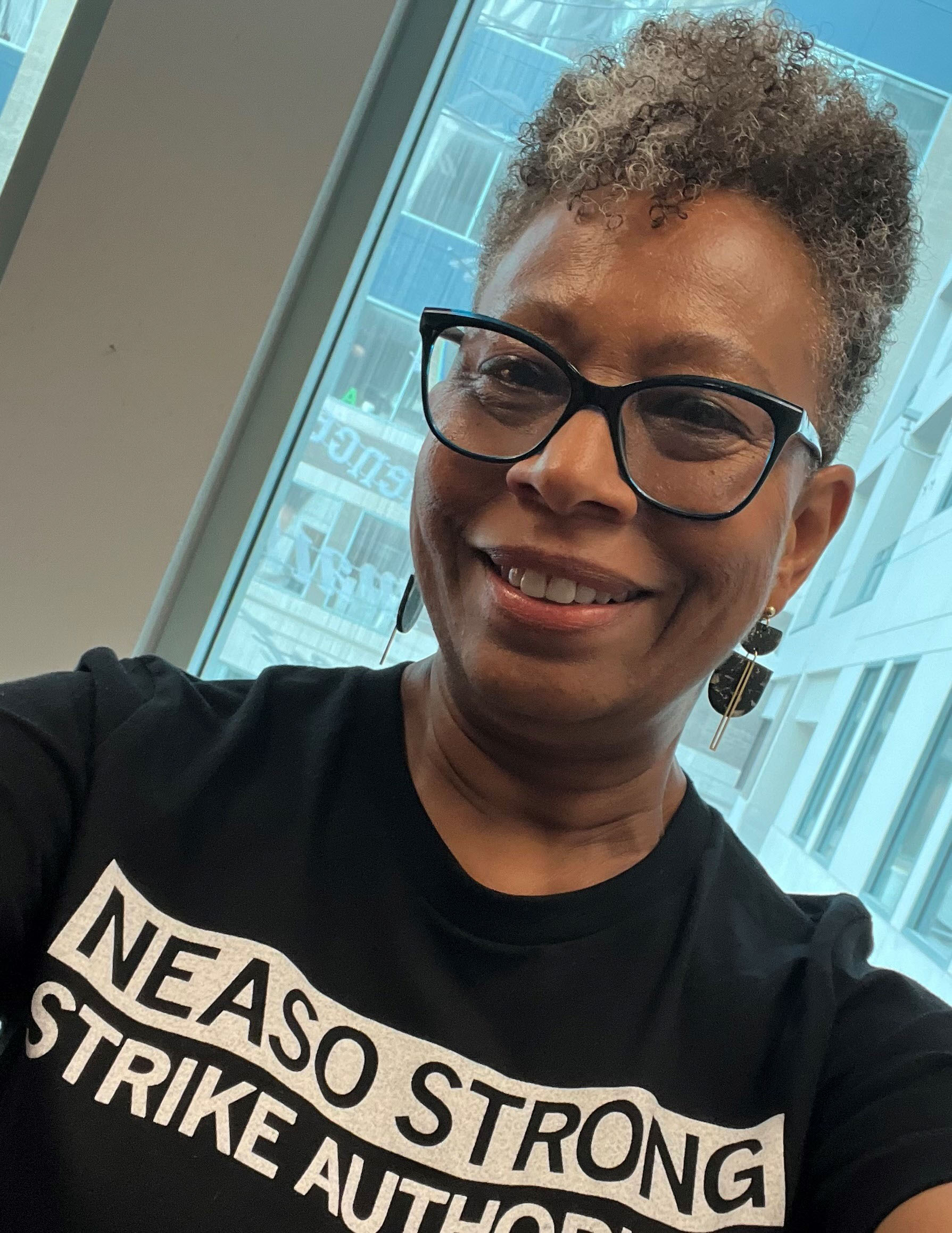 A woman wearing glasses and a t-shirt that reads "NEA SO STRONG"