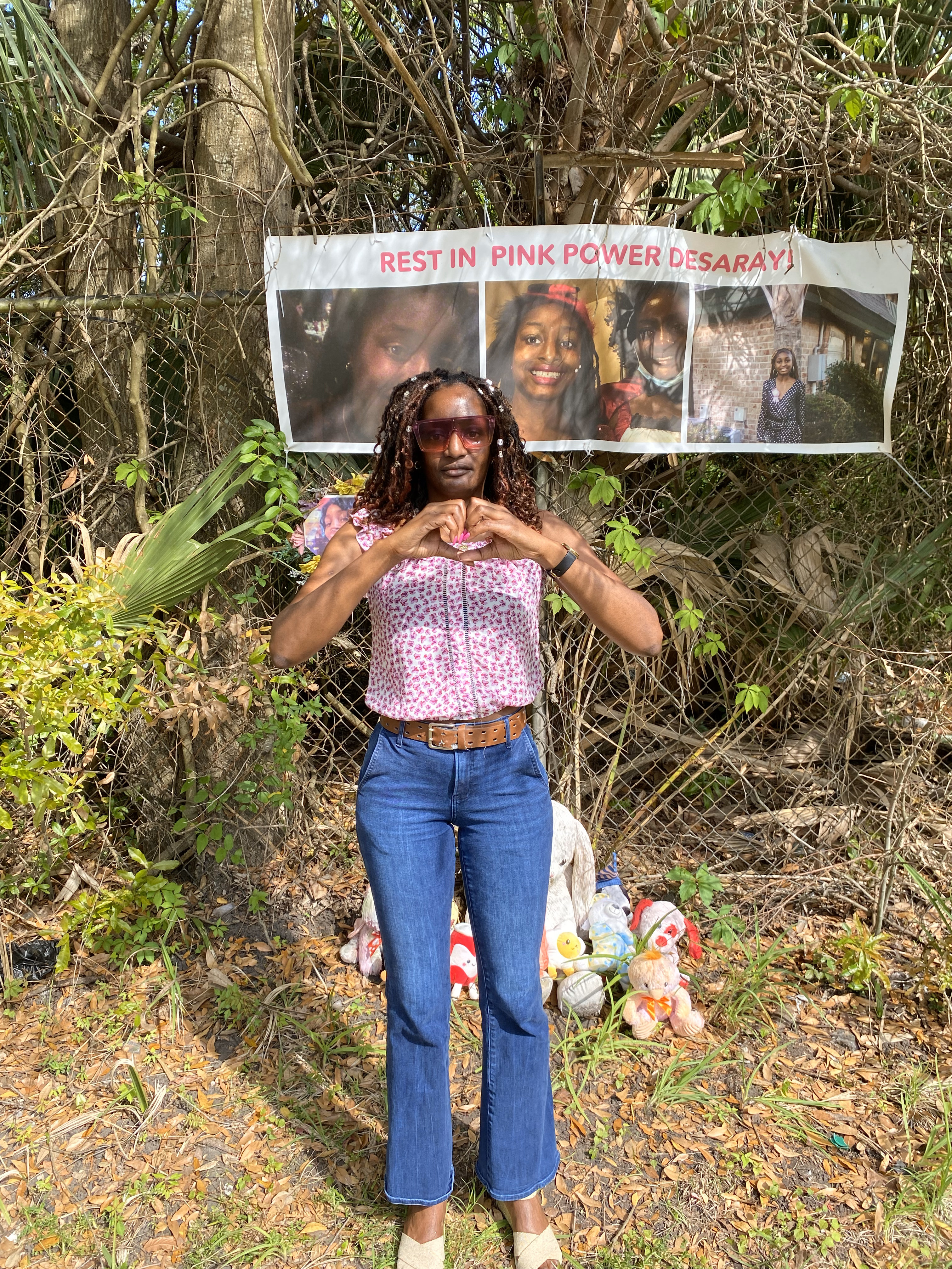 A woman in a pink and white short-sleeve shirt and jeans and holding her hands in the shape of a heart stands in front of a fence where a banner hangs. The banner has three photos of a teenage girl on it and the words "Rest in Pink Power Desaray". There are several stuffed animals on the ground behind the woman, in front of the fence.