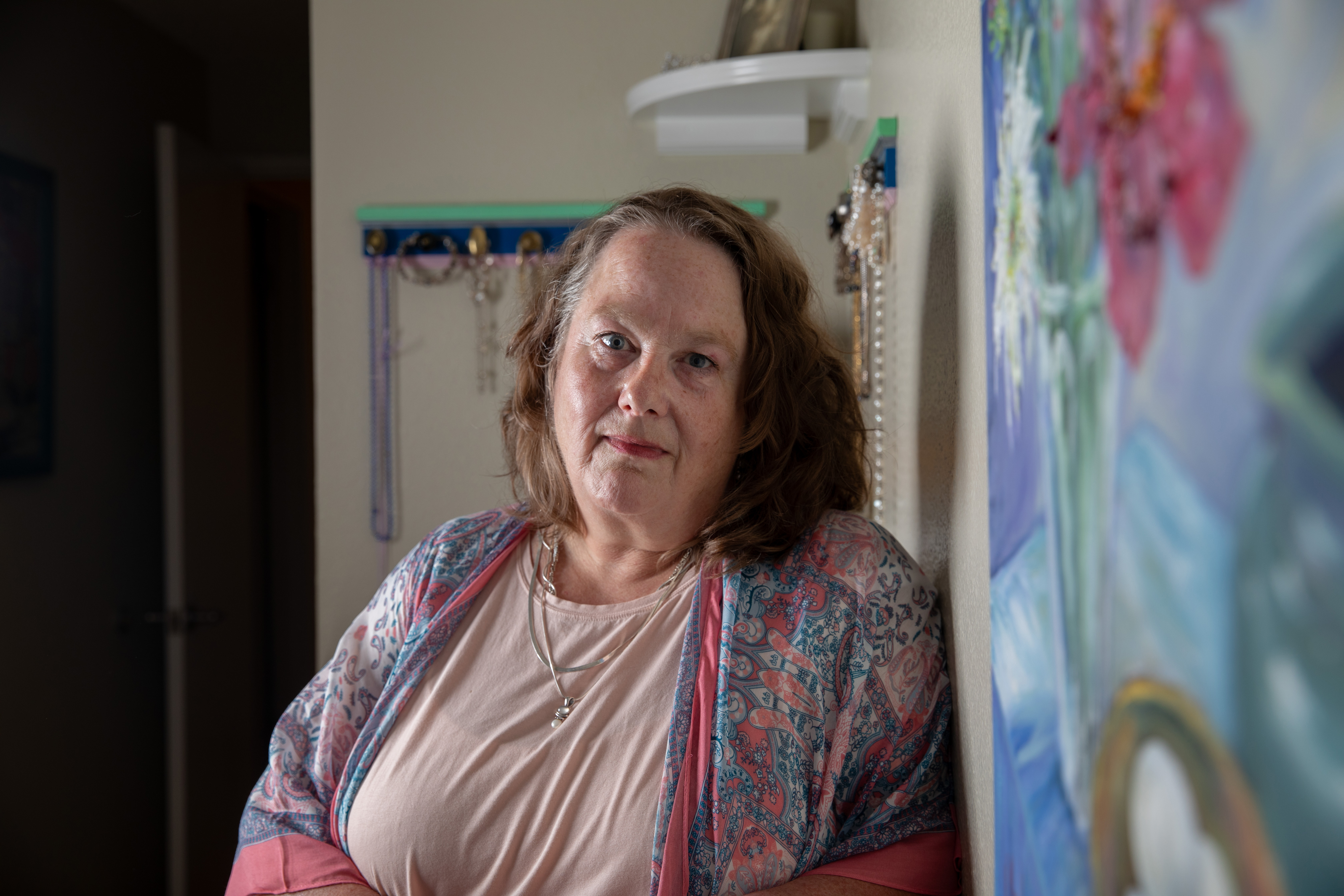 A woman in a pink tshirt and paisley cardigan leans against a wall