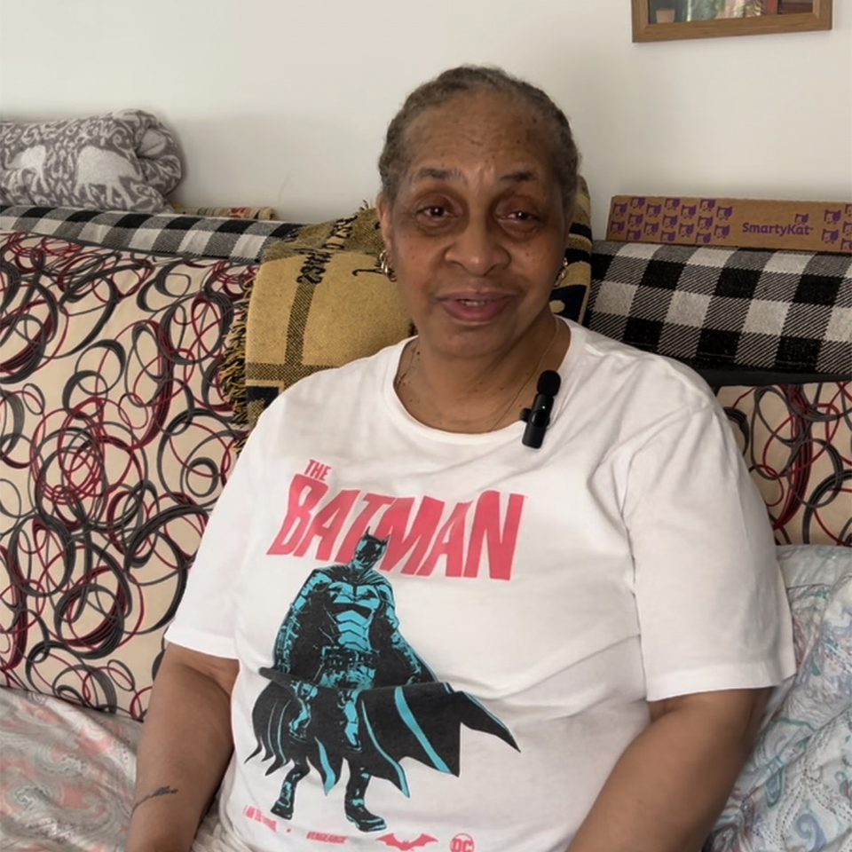 An senior woman wearing a Batman t-shirt sits on a couch and looks at the camera and smiles gently.