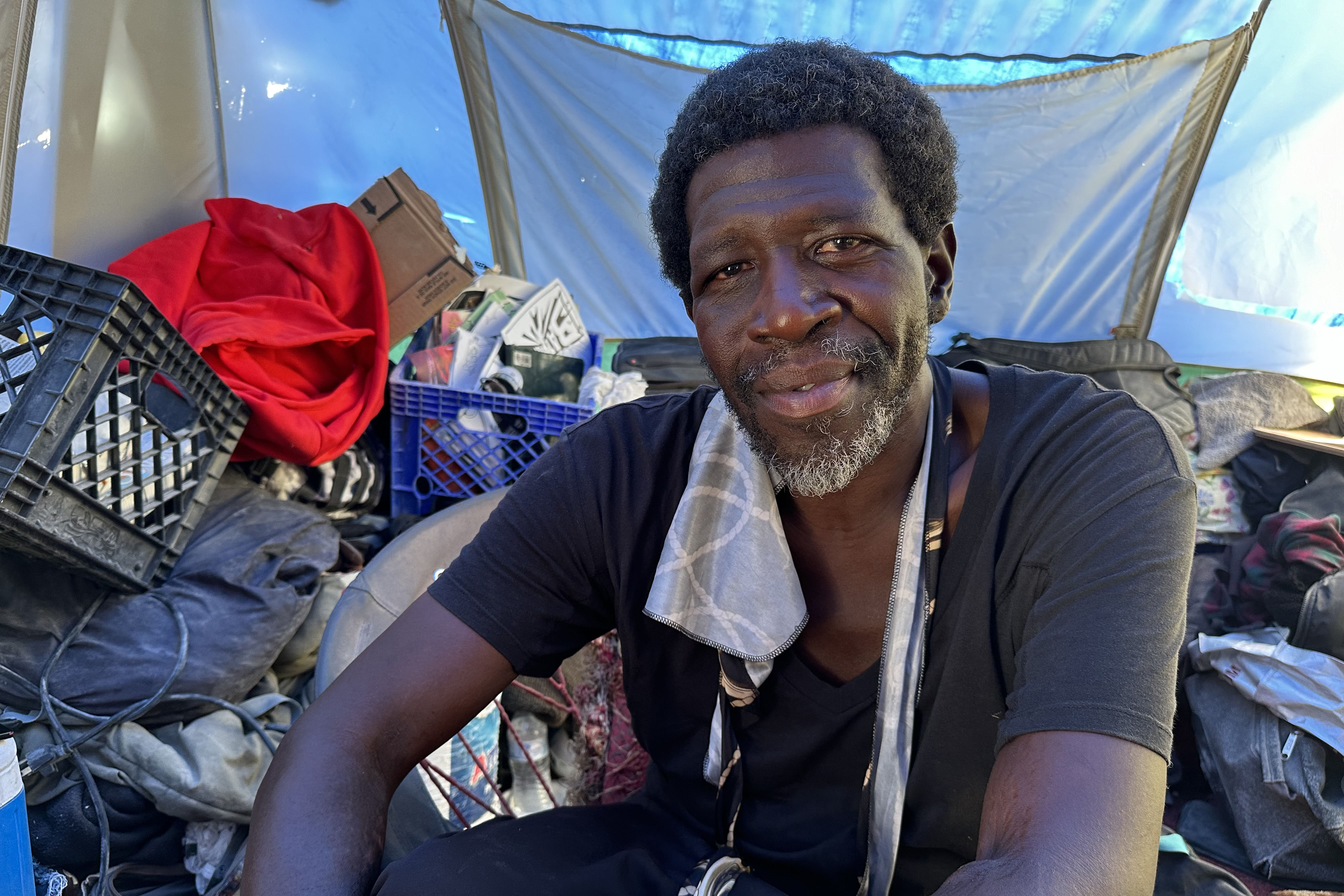 A portrait of Maurice Clark, who is sitting amongst his belongings. He has a warm expression as he looks towards the camera.