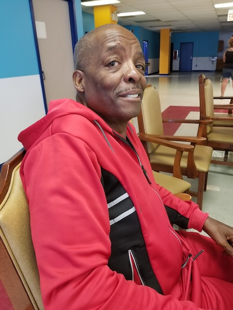 A senior man in a red and black zip up shirt sits on a chair and looks at the camera