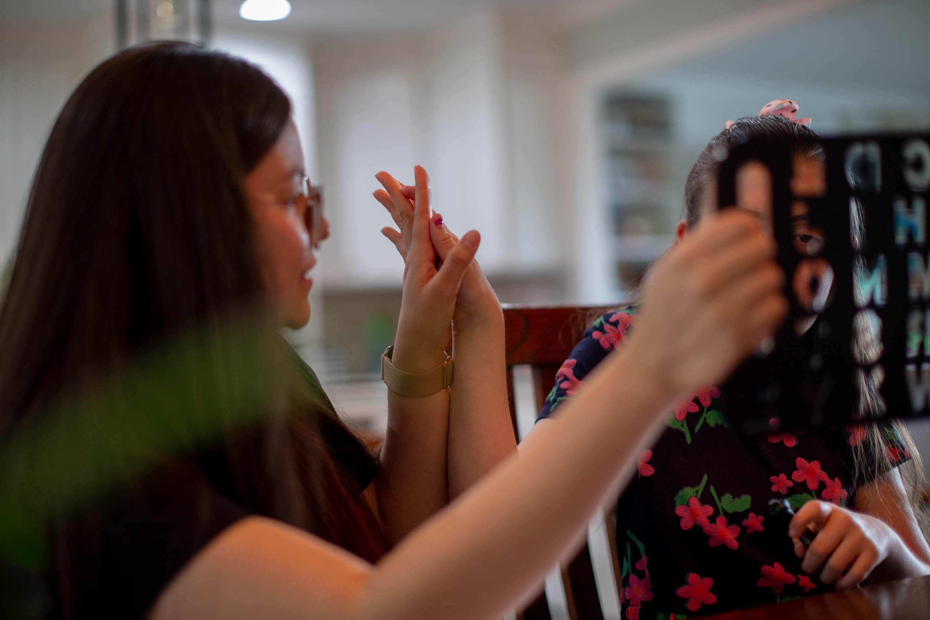 A woman with long brown hair wearing glasses holds the hand of a girl as she helps her with homework