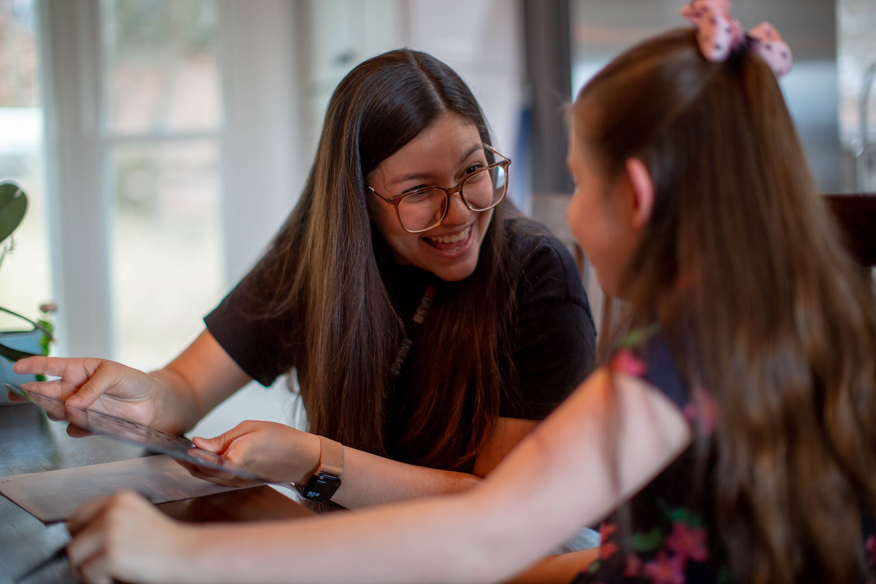 A woman with long brown hair wearing glasses smiles at a girl wearing a pink bow in her hair