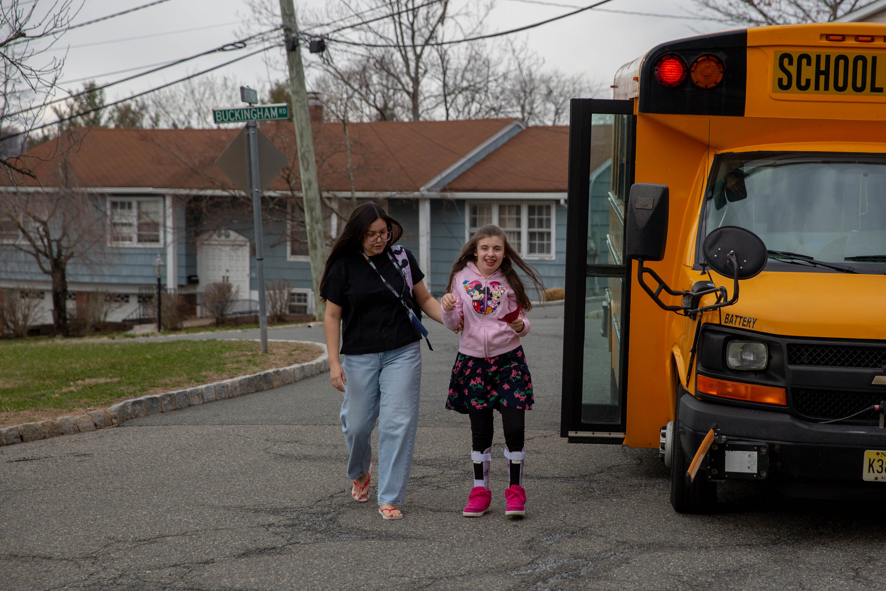 A woman with long brown hair wearing glasses walks away from a school bus with a girl wearing a pink sweatshirt and flowered dress