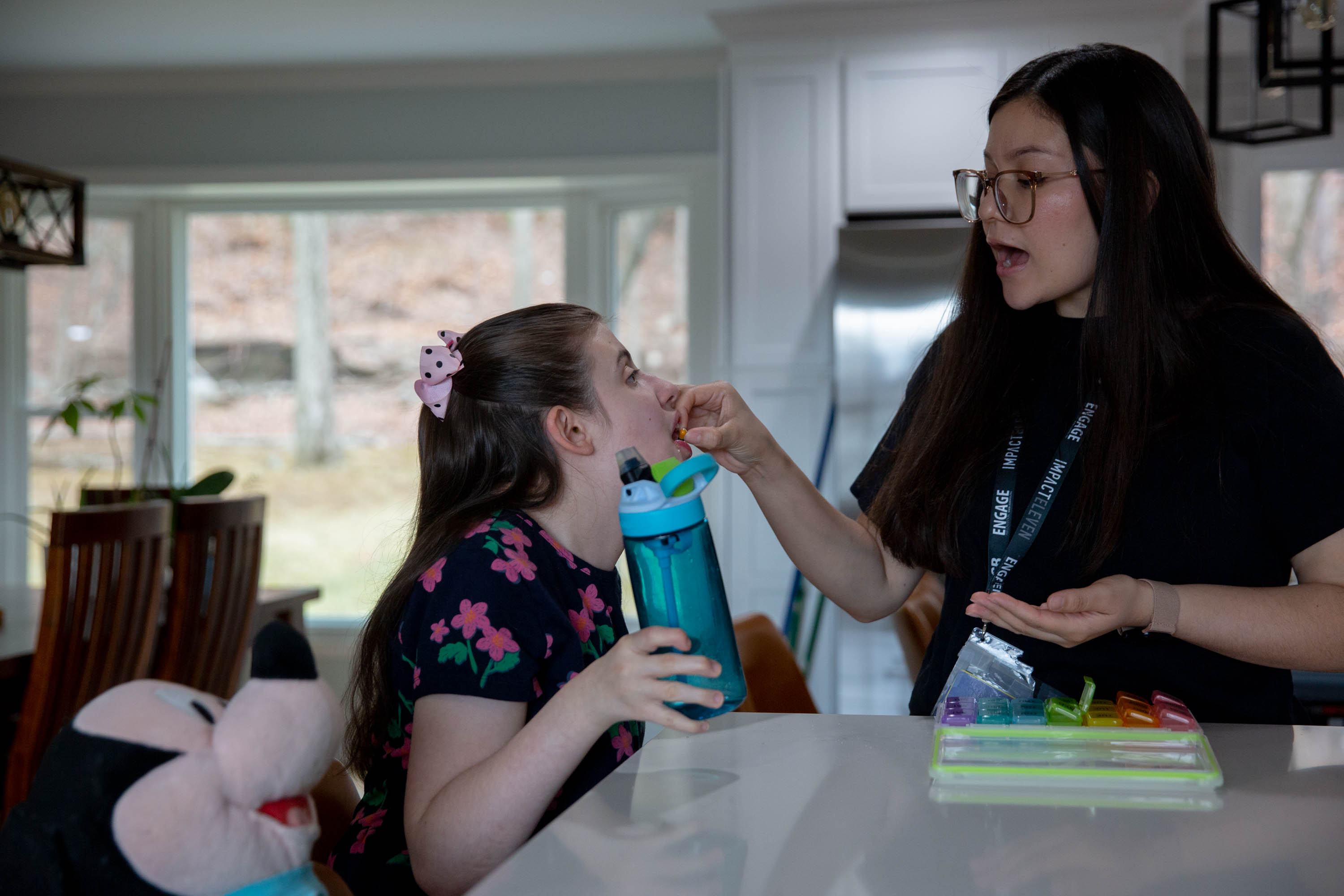 A woman with long brown hair wearing glasses puts a pill into the mouth of a girl sitting at a countertop with her mouth open
