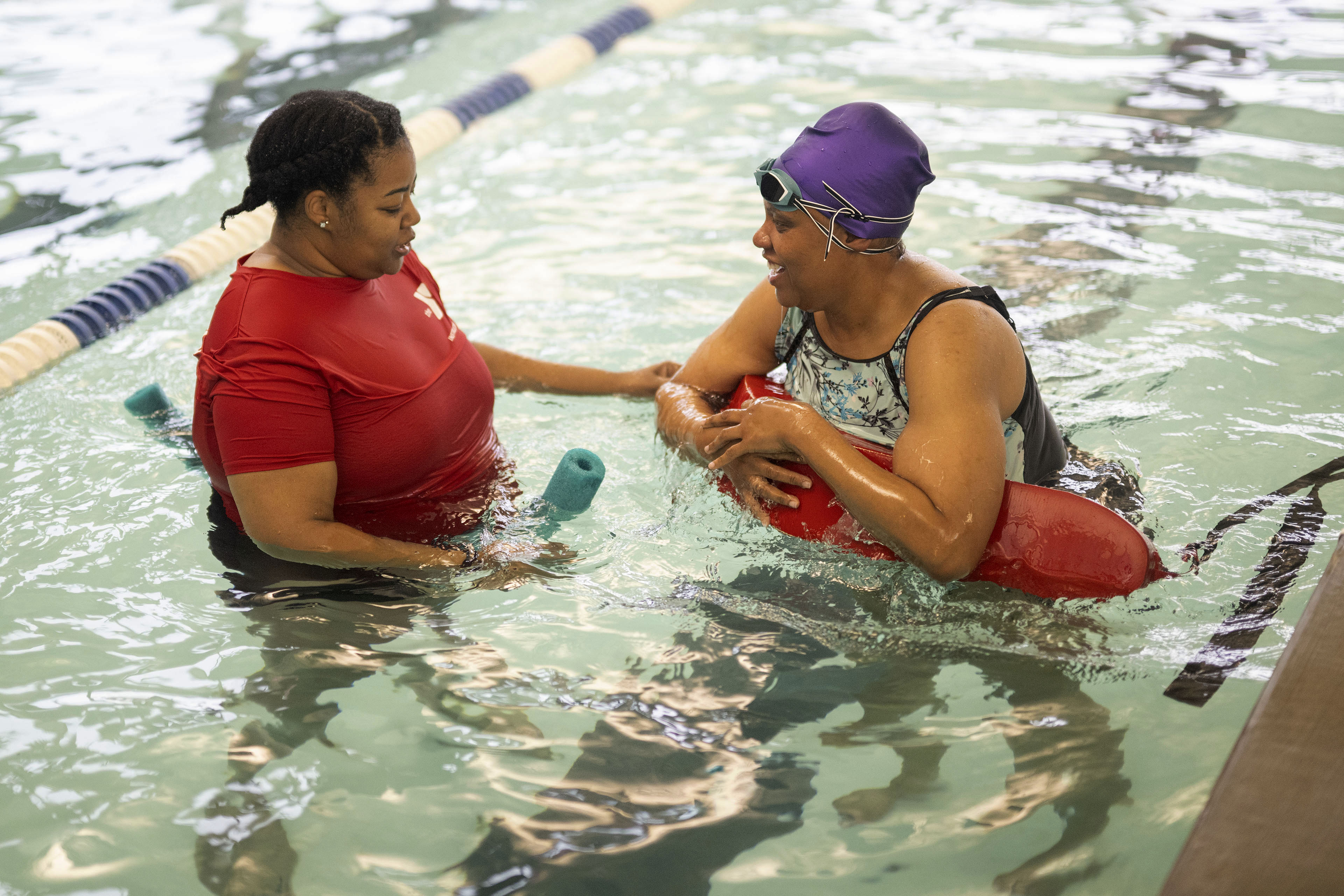 A swim instructor talks with a woman wearing a purple swim cap and goggles as she holds a red floatation device in a pool