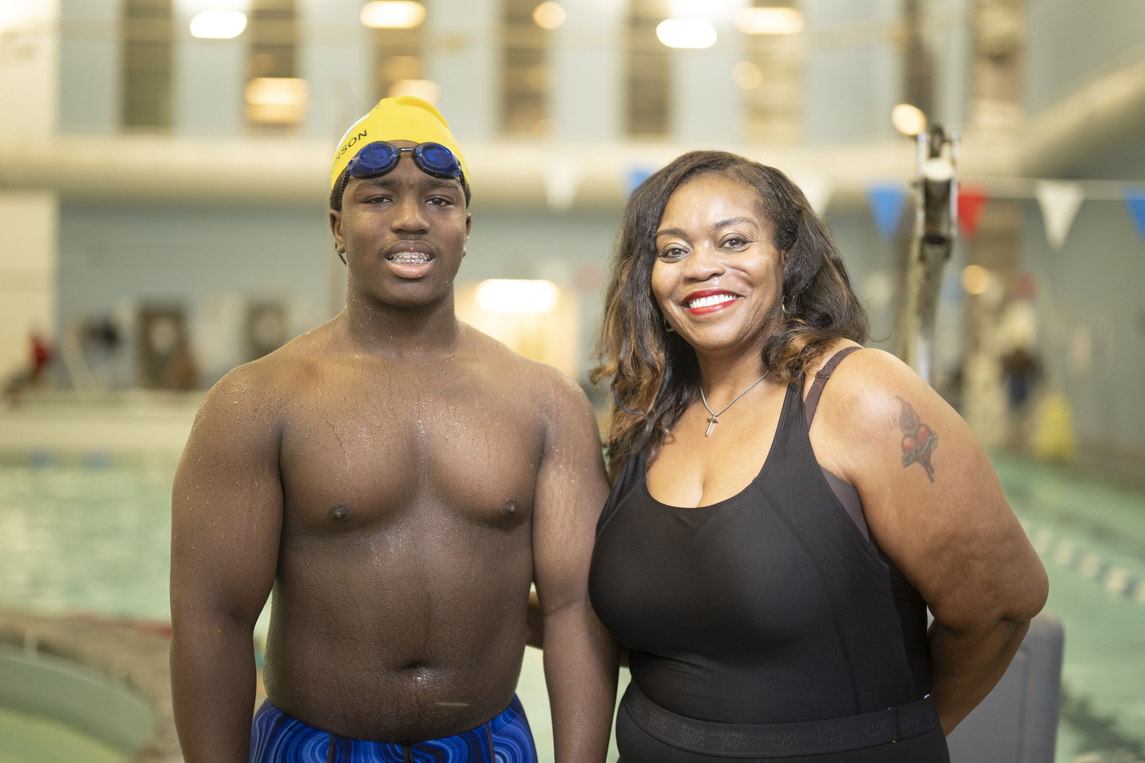 A young man wearing a yellow swim cap, goggles, and blue swim shorts poses for a photo next to a woman in a black bathing suit